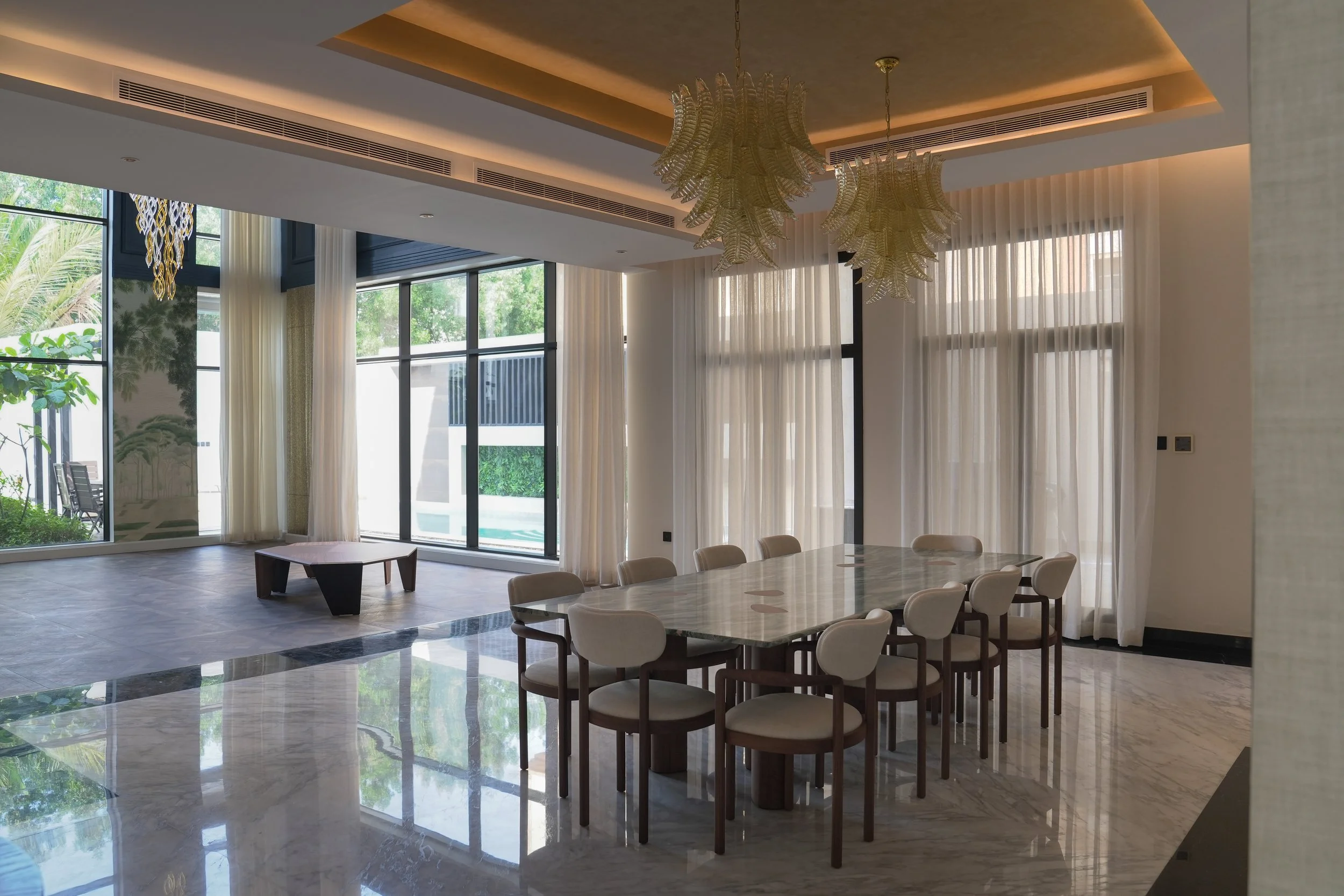 A spacious, well-lit dining area with a marble dining table, eight beige chairs, and large windows with sheer curtains, overlooking a garden and outdoor seating.