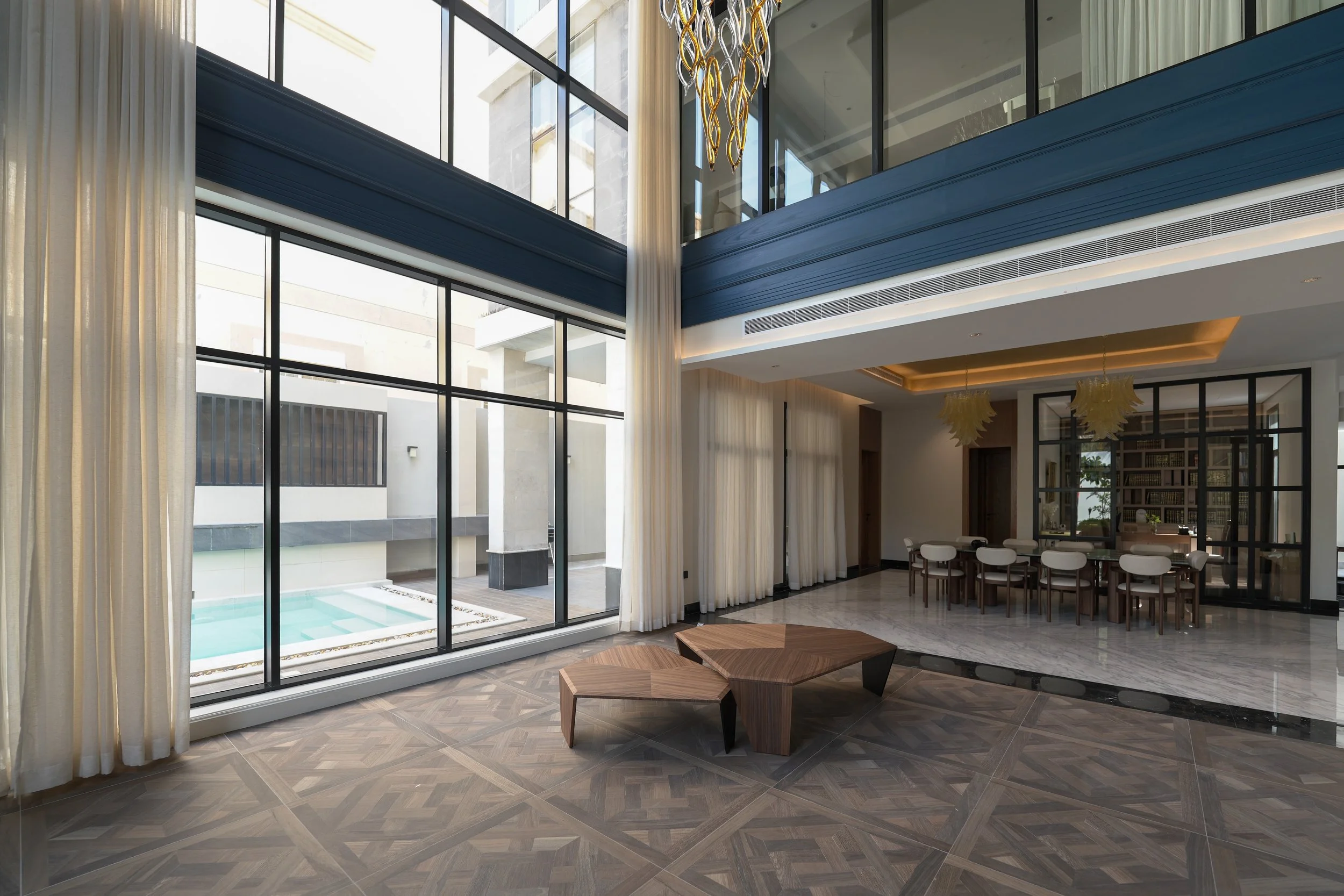 Modern spacious living room with large floor-to-ceiling windows, a pool outside, contemporary wooden coffee table, and a dining area with chairs and pendant lights.