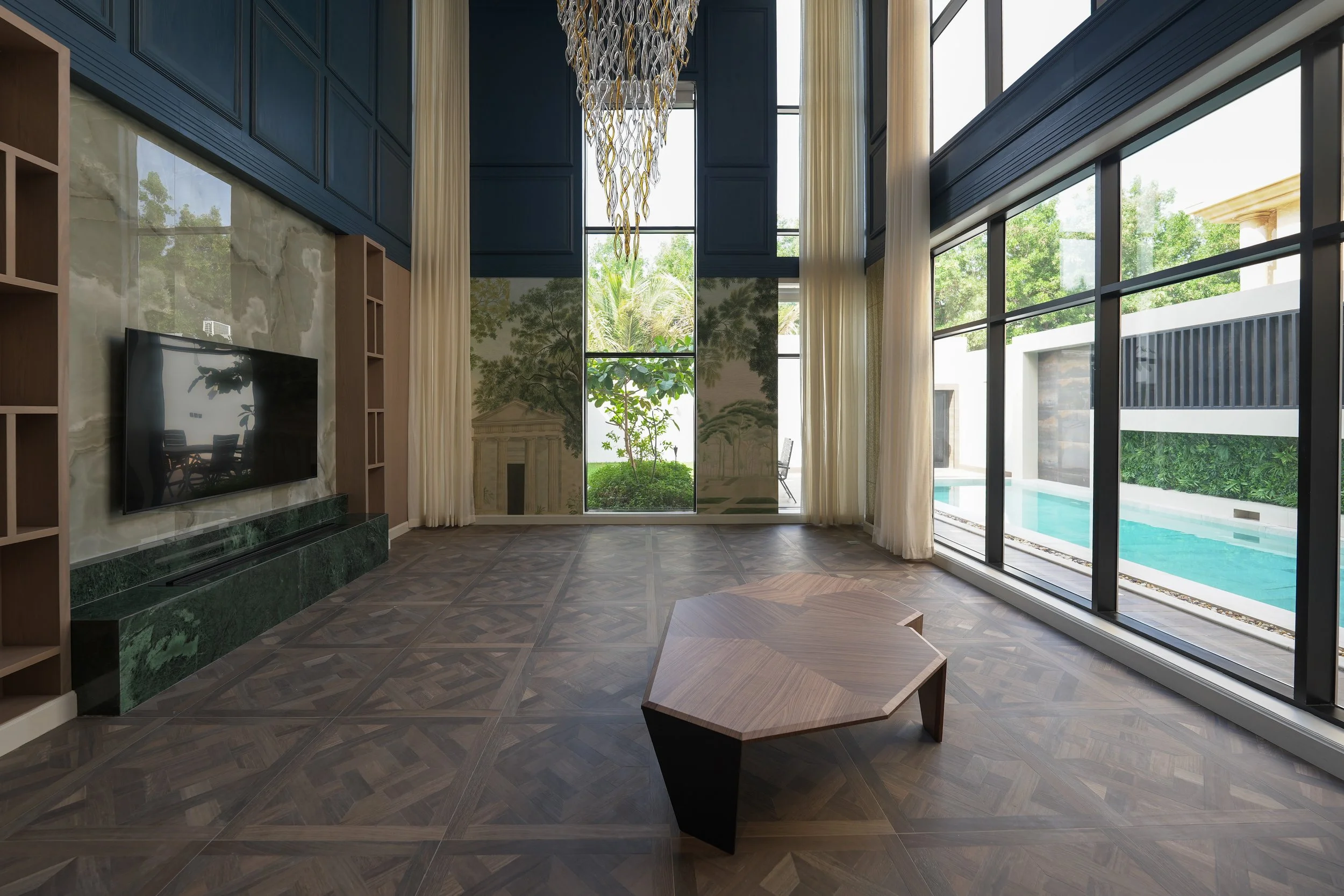 Modern living room with large floor-to-ceiling windows, a wall-mounted TV, a decorative chandelier, and a view of a pool and outdoor greenery.