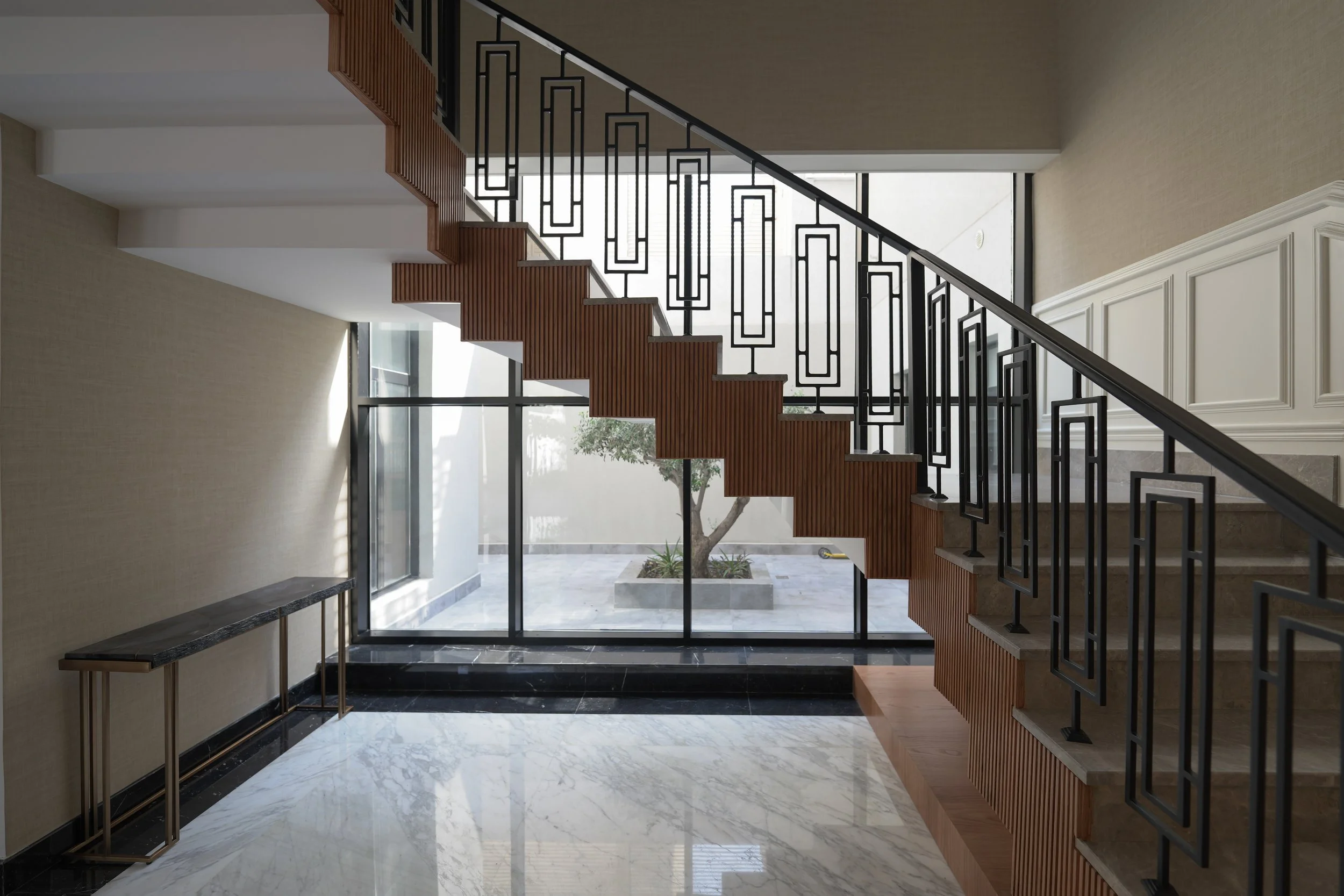 Interior view of a modern staircase with black metal railings and wooden steps, leading to an upper floor, with a large window and a small tree outside.