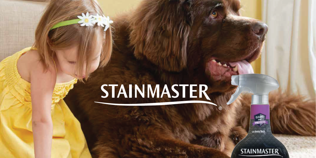 A young girl wearing a yellow dress and a daisy flower headband sitting next to a large brown dog indoors. A bottle of StainMaster pet stain remover spray is in the foreground.