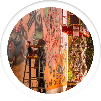 A person on a ladder painting a large vibrant mural on a wall, with a cherry picker nearby.