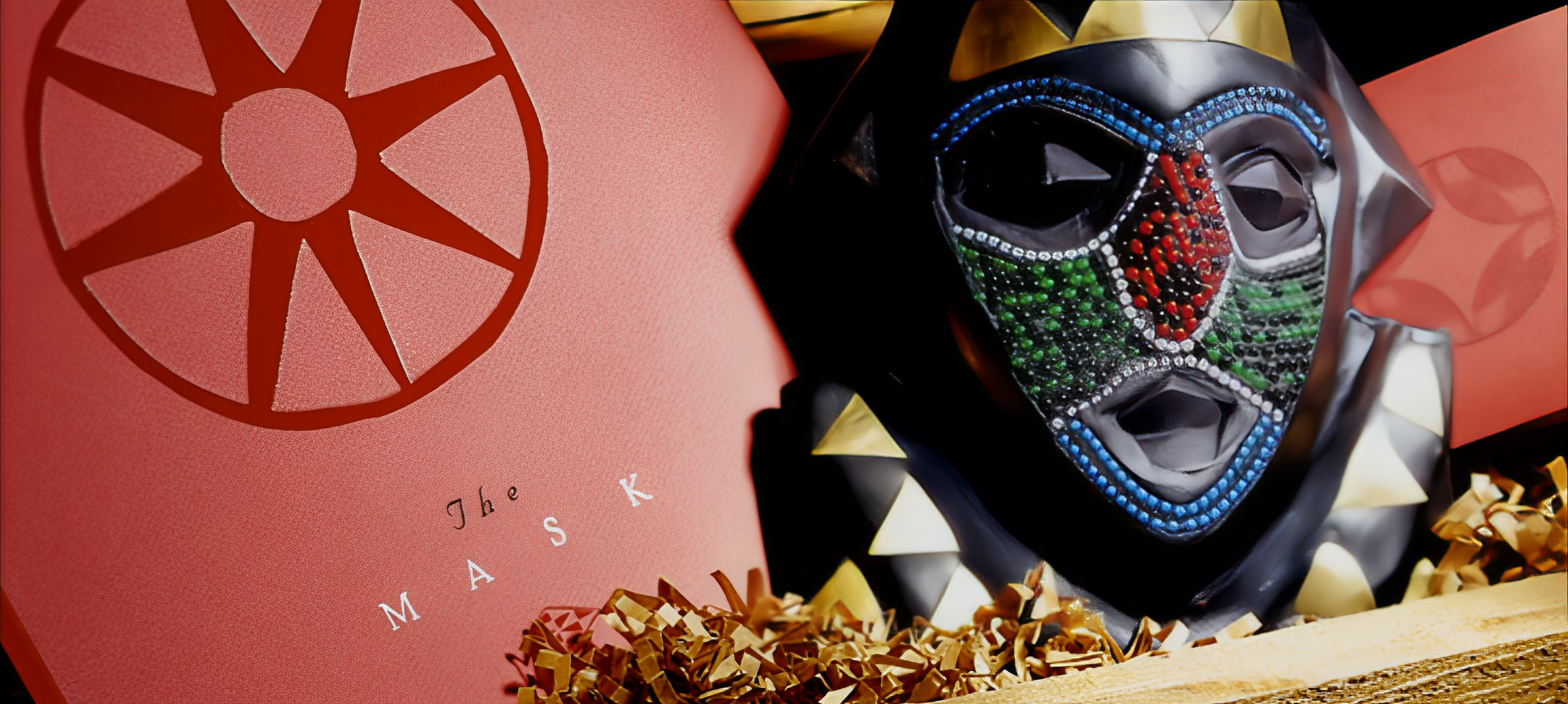 Pink box with red citrus slice graphic and the words "The MASK" in black, next to a colorful decorative mask with gemstones, gold spikes, and a mix of green, red, blue, and black accents, surrounded by shredded paper.