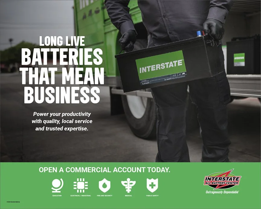 A person in black clothing and gloves holding a black Interstate battery, standing near a green outdoor equipment truck. The image includes text promoting long-lasting batteries for business, with a call to open a commercial account, featuring icons for education, electrical, fire safety, medical, and public safety, and the Interstate logo.