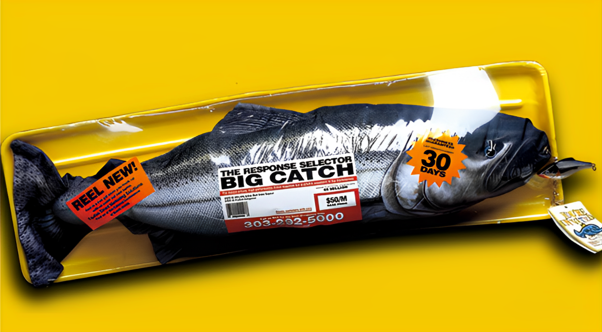 A robotic fish with a fishing lure in its mouth, placed on a yellow surface, with a label reading "BIG CATCH" and advertising "30 days" for a fishing reel brand.