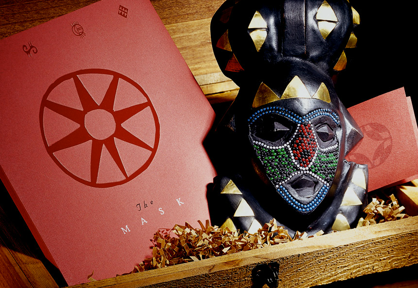 Decorative African mask with colorful beadwork, in a wooden box next to a pink card with a red circle logo and the words "The Mask" on it, surrounded by crinkle paper.