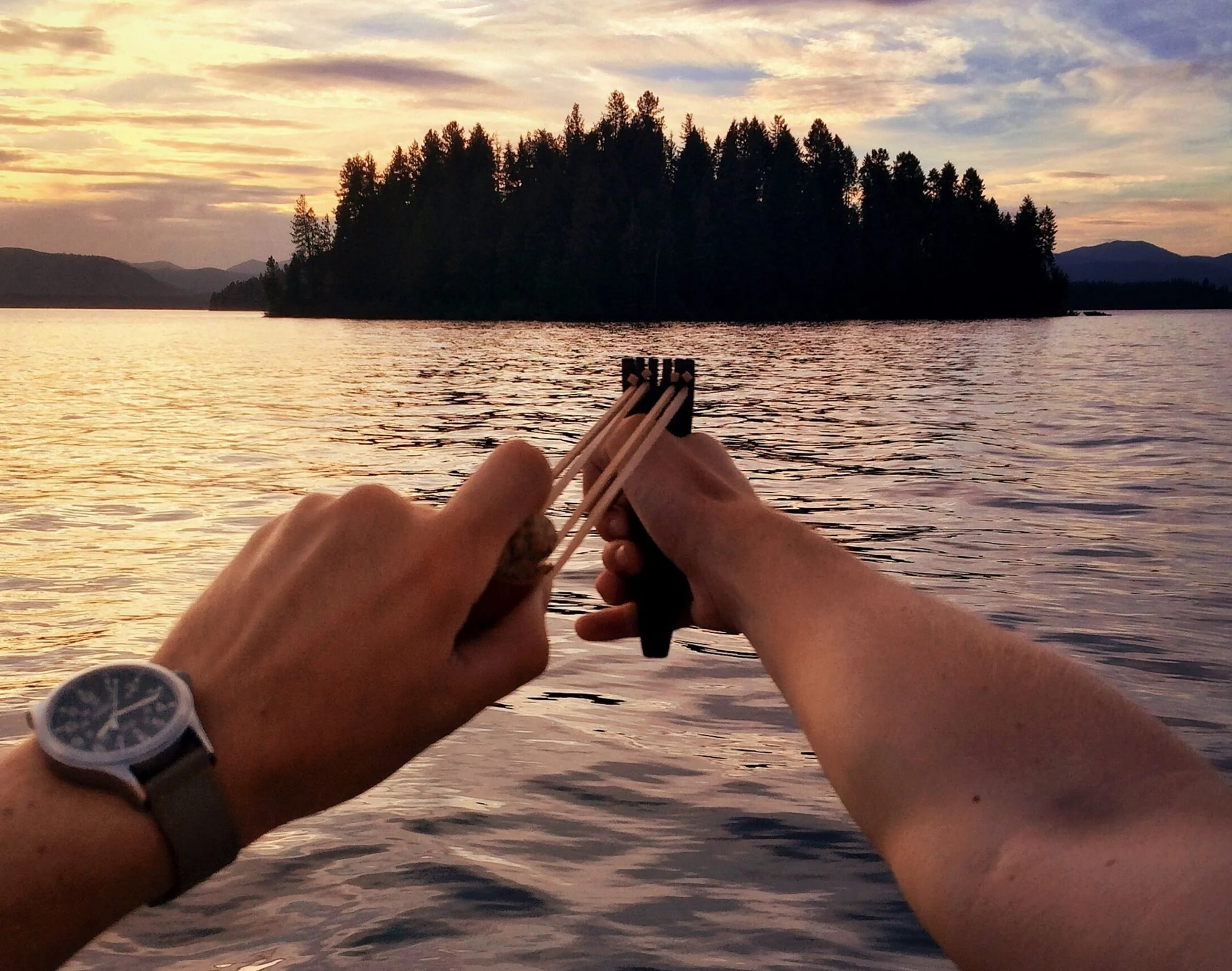Two hands holding a slingshot over a body of water during sunset, with a wooded island and mountains in the background.