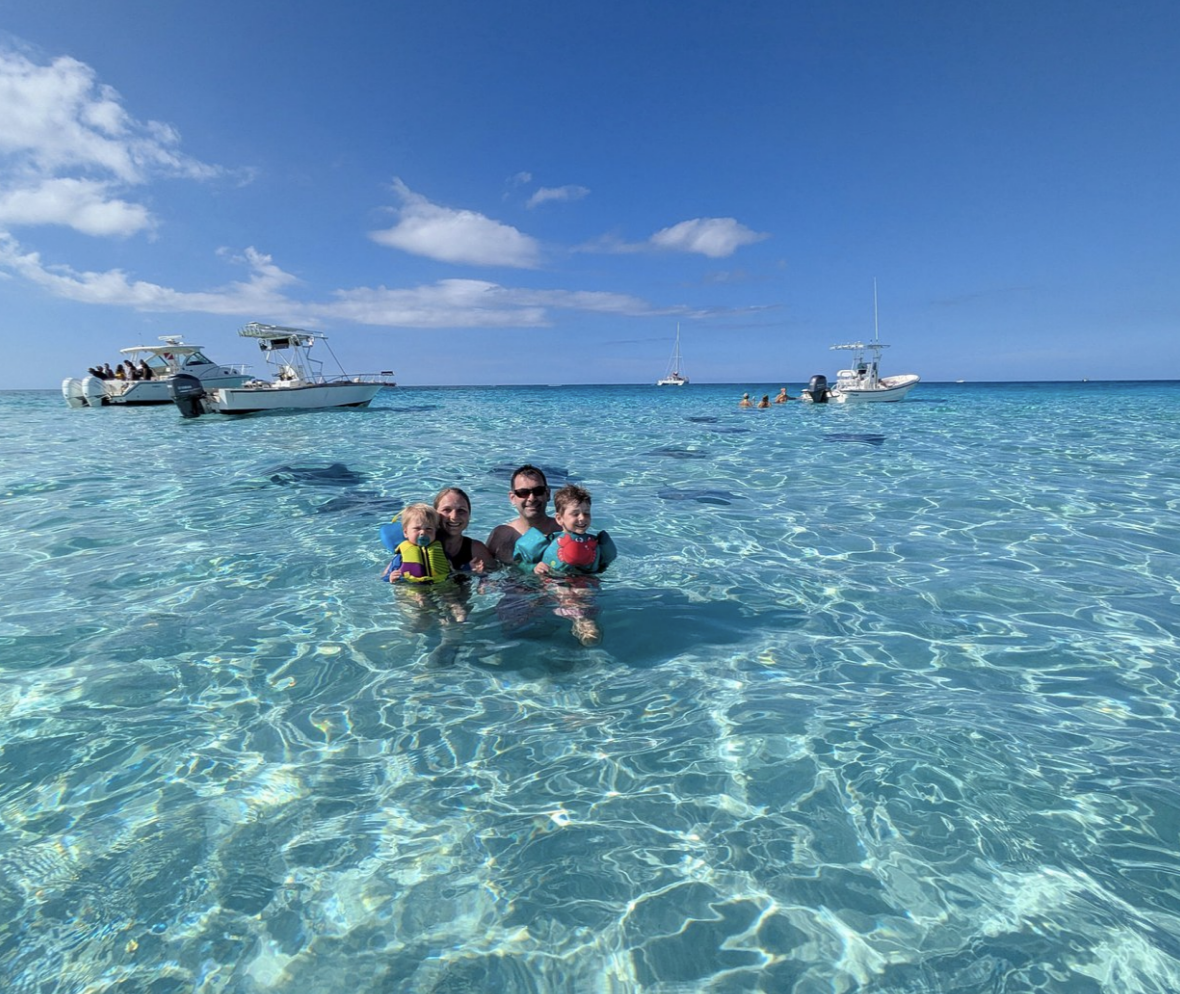 Adults and children enjoying clear shallow water with boats in the distance.