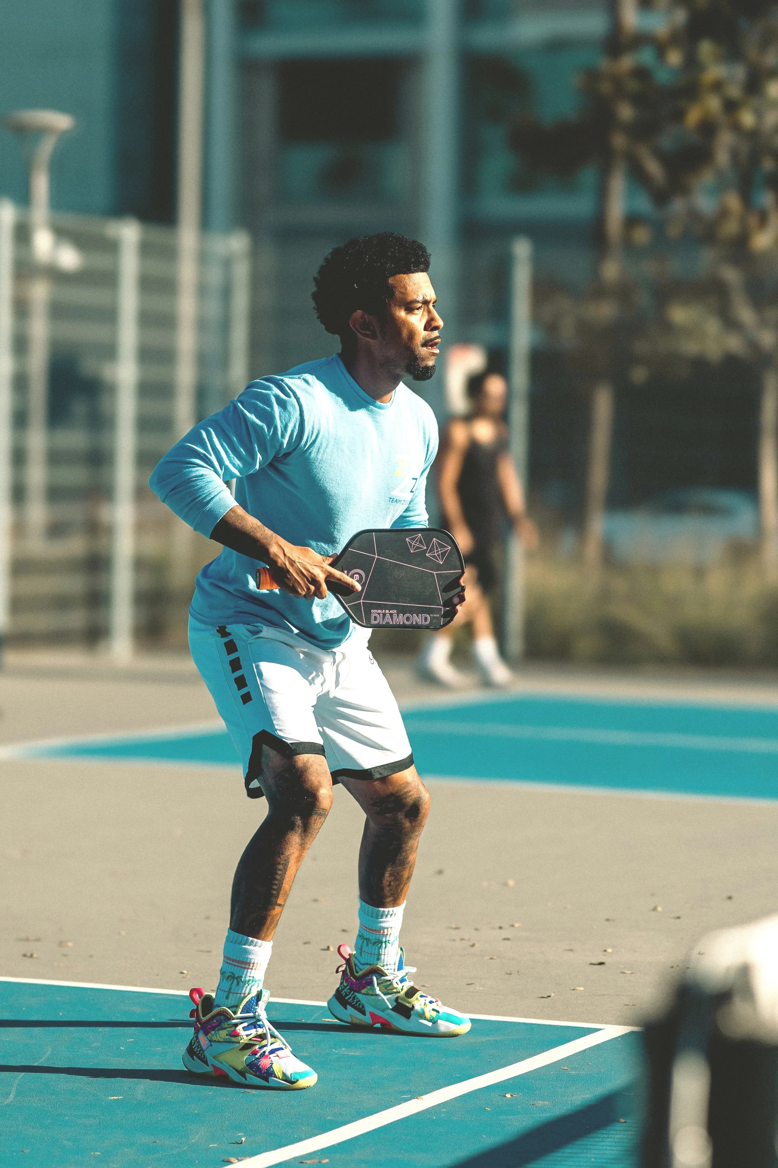 A man is playing pickleball on an outdoor court, holding a paddle in his right hand, wearing a blue long-sleeve shirt, white shorts, colorful sneakers, and sports socks.