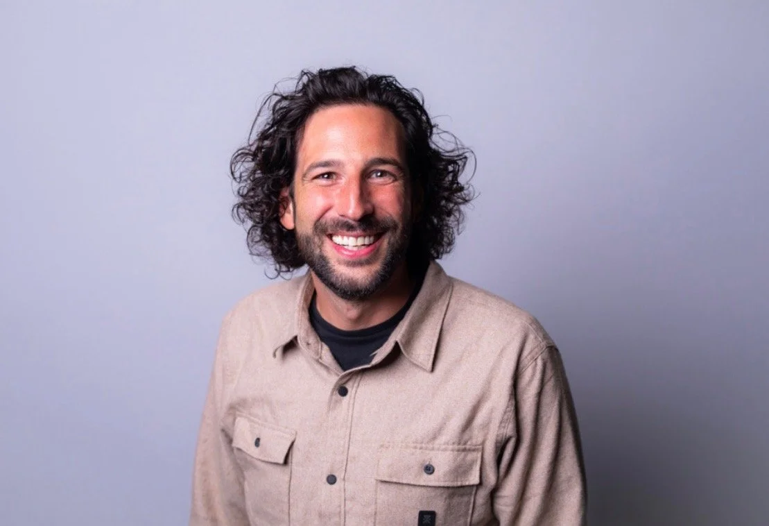 Man with curly dark hair and beard smiling, wearing a beige jacket and black shirt, against a light gray background.