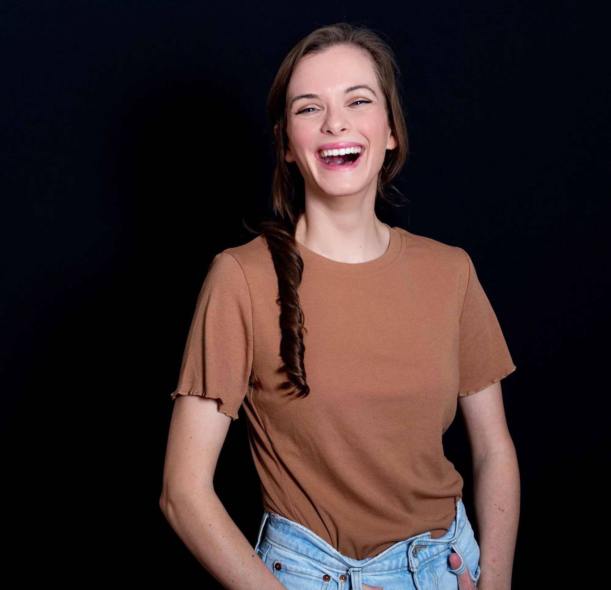 A woman with long brown hair in a loose braid, smiling and laughing, standing against a black background, wearing a brown T-shirt and light denim shorts.
