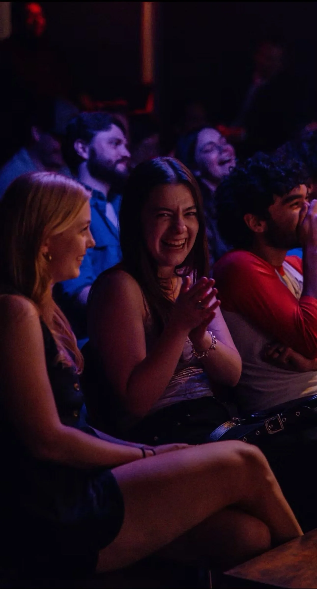 People sitting in an audience, laughing and smiling at a comedy show or performance, under dim, colorful stage lighting.