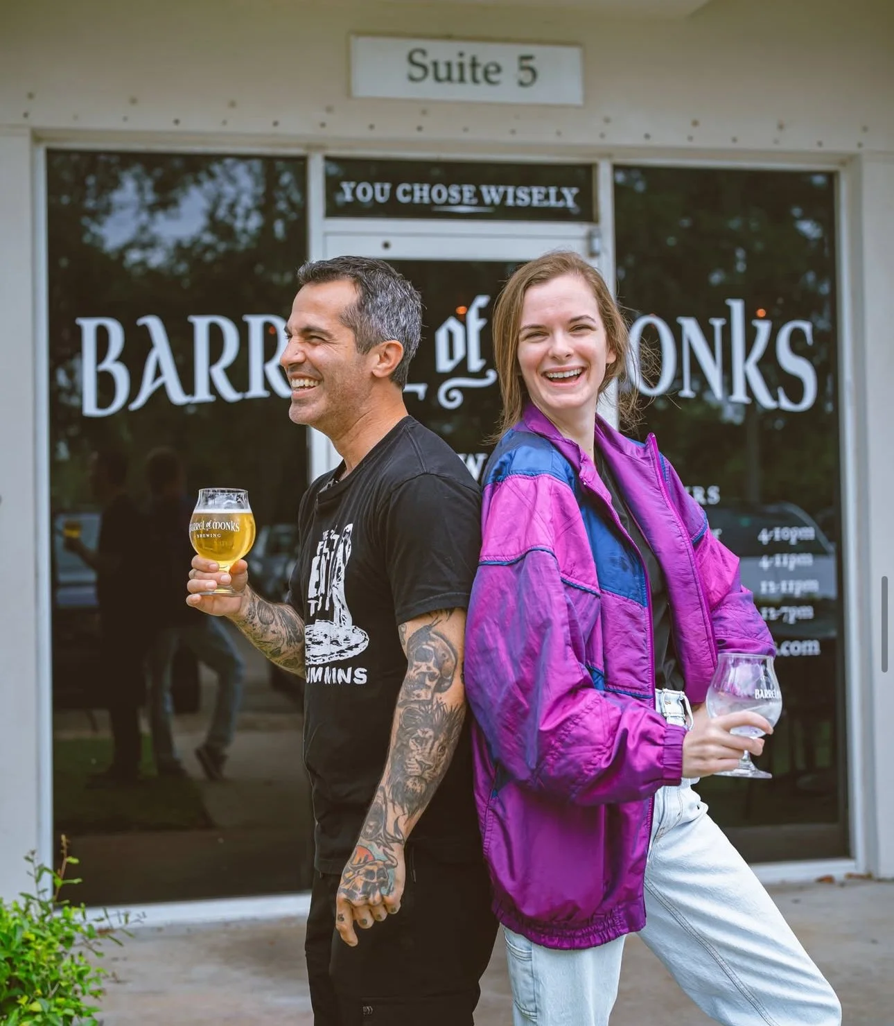 A man and woman standing back to back outside a bar called Barr of Monks, each holding a wine glass with beer, smiling and enjoying their time.