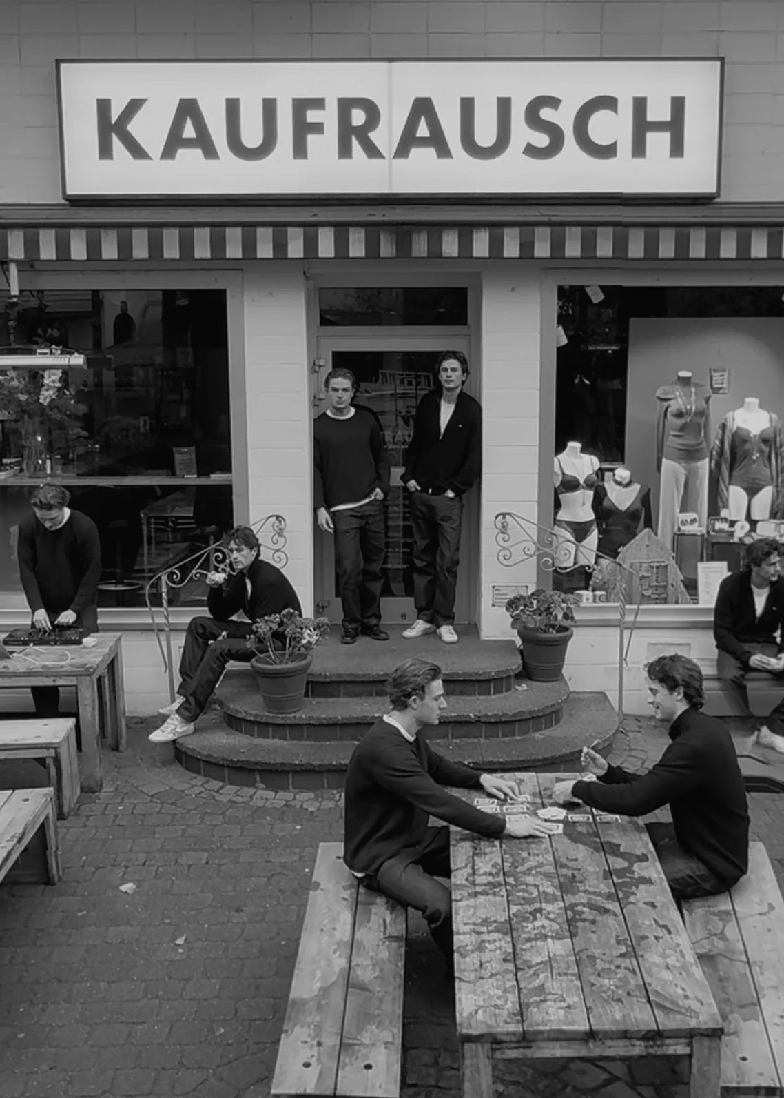 Schwarz-Weiß-Foto von einer Straßenszene vor einem Geschäft mit dem Schild "KAUFRUCSCH". Es zeigt mehrere Personen, einige sitzen an Tischen, andere stehen vor dem Geschäftseingang, während zwei junge Männer in dunklen Jacken auf der Treppe vor dem Laden stehen.
