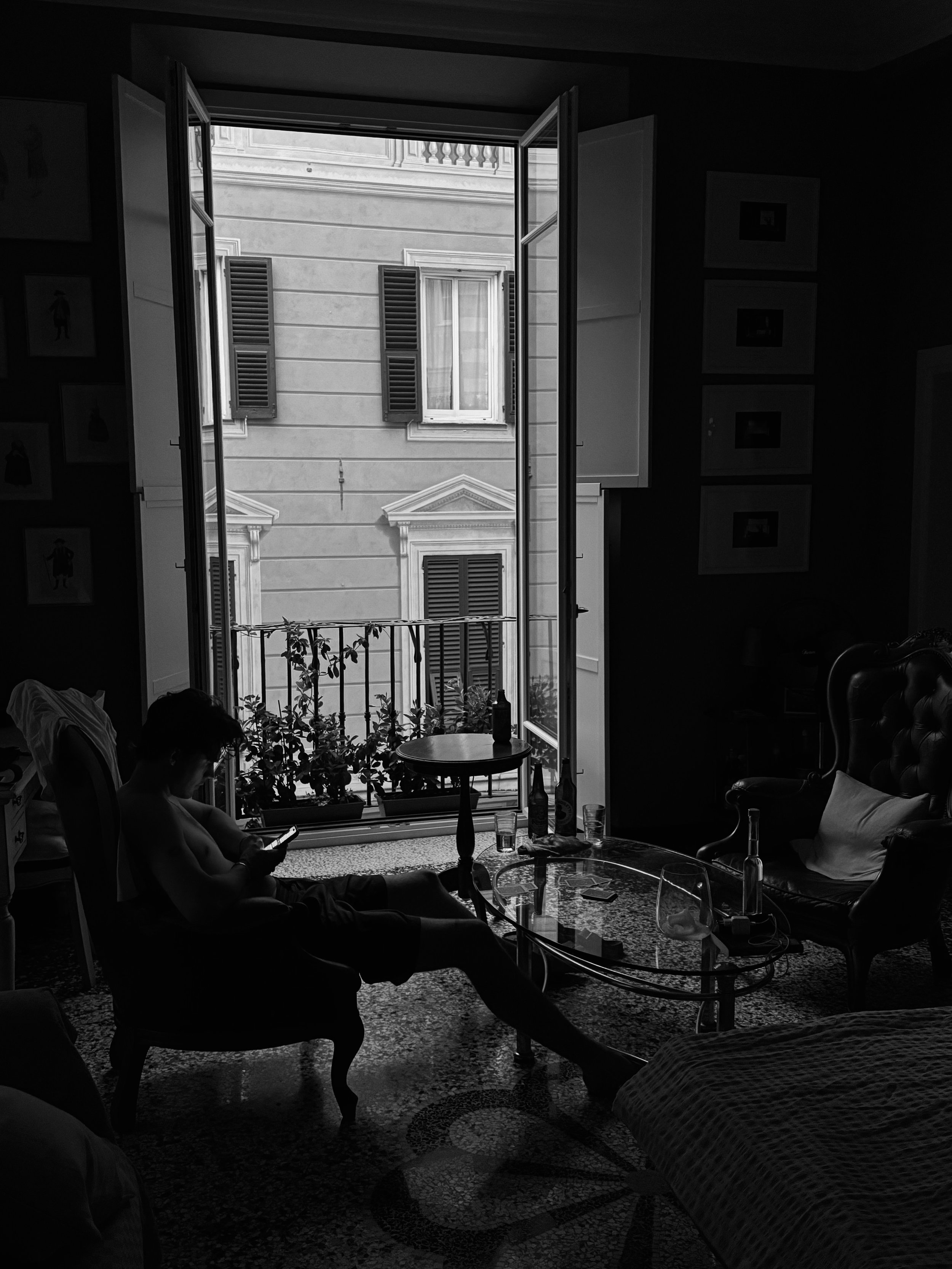 Ein dunkel gehaltenes Wohnzimmer mit offenen französischen Fenstern, durch die Tageslicht strömt. Es gibt eine Person, die auf einem Vintage-Sessel sitzt und ein Smartphone benutzt, und einen Couchtisch mit Getränkeflaschen, einem Glas und anderen Gegenständen. Neben dem Fenster befinden sich Pflanzen im Balkon. Im Hintergrund sind die Fassade eines Gebäudes mit erkennbaren Fenstern und Shutters sichtbar.