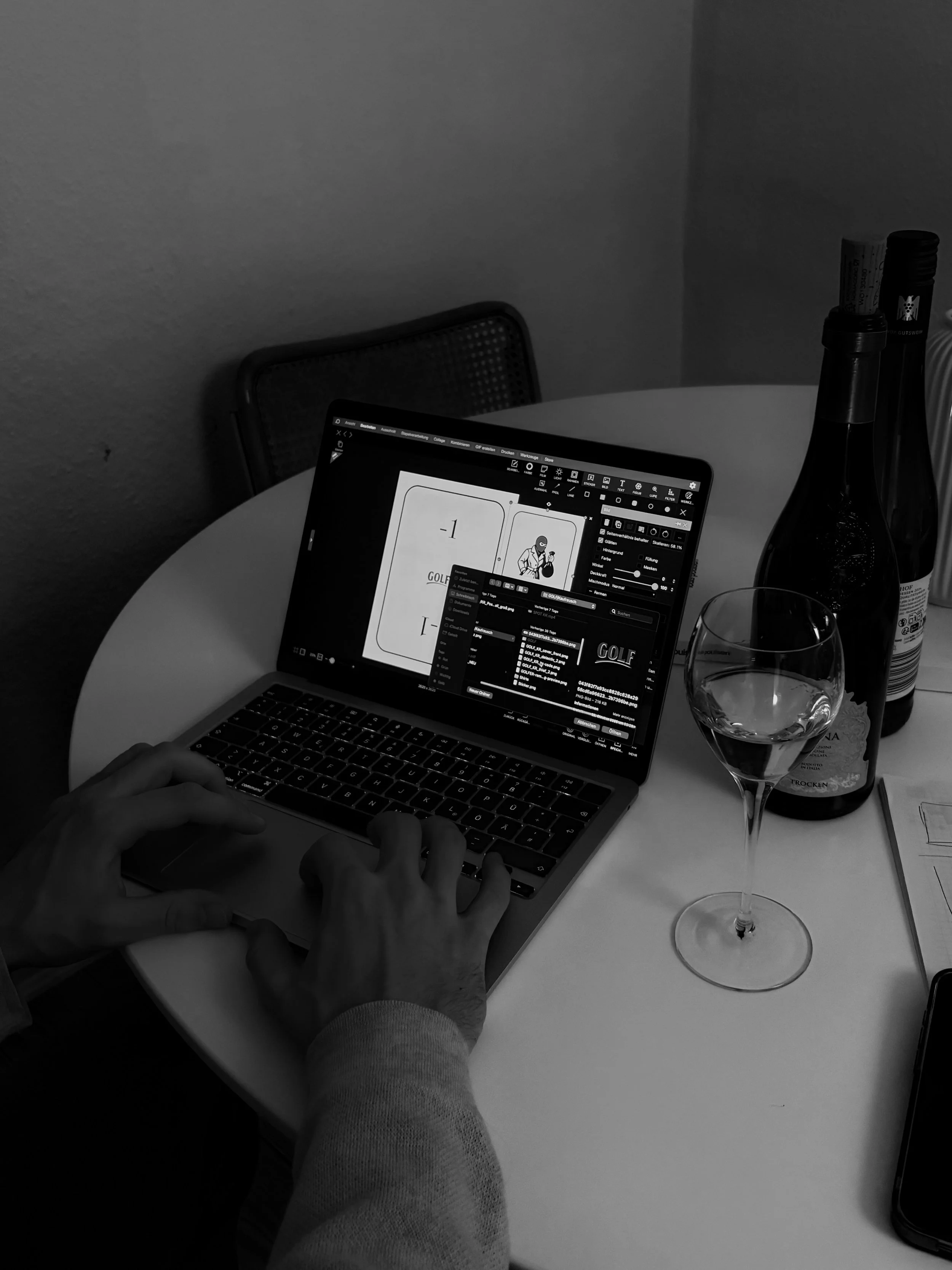 Schwarz-weiß Foto eines Laptops auf einem Tisch, auf dem eine Person arbeitet, mit einer Hand auf der Tastatur. Es gibt auch eine Weinflasche, ein Weinglas mit Wasser, und einige Papiere auf dem Tisch.