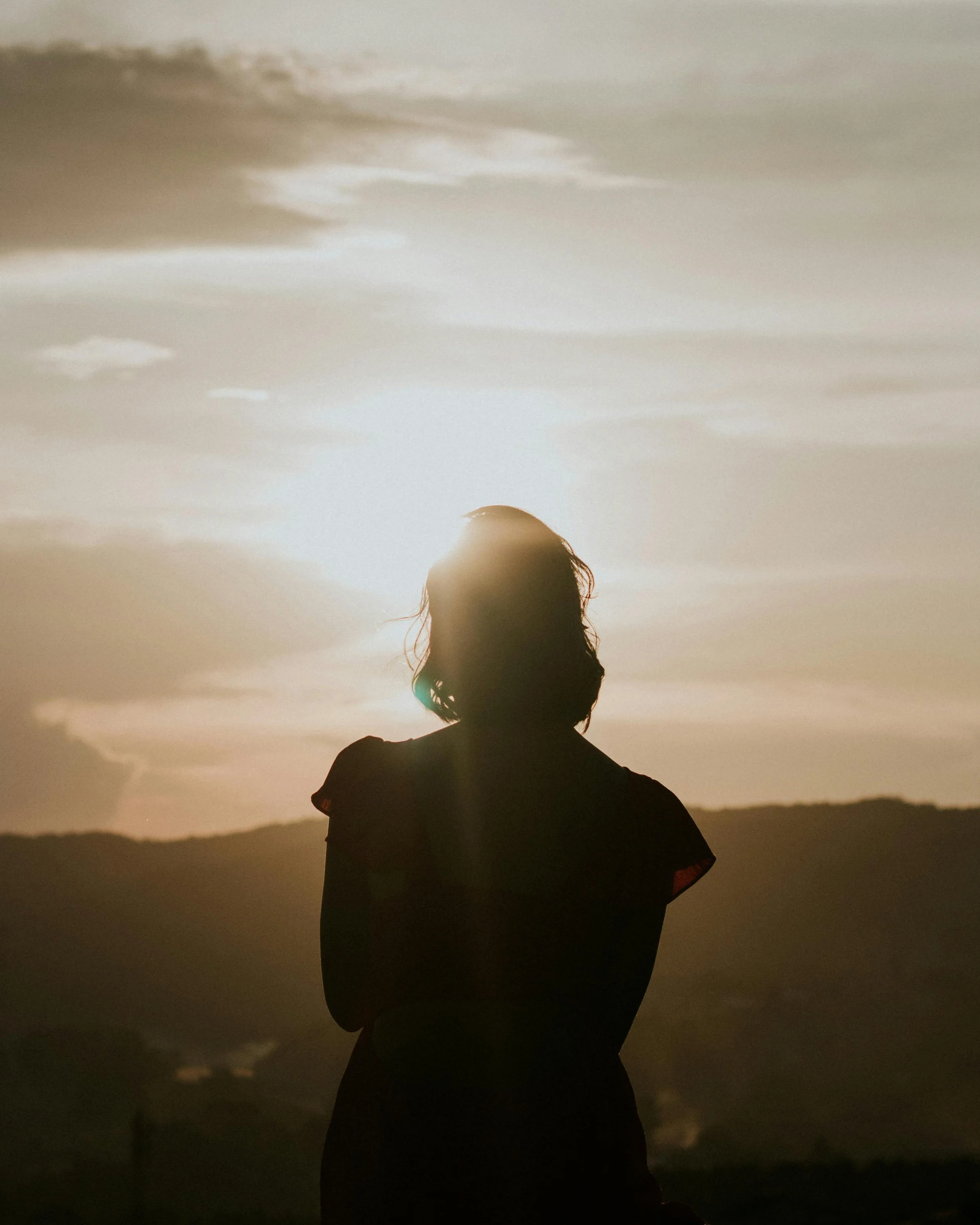 Silhouette of a woman standing outdoors during sunset or sunrise with mountains in the background.