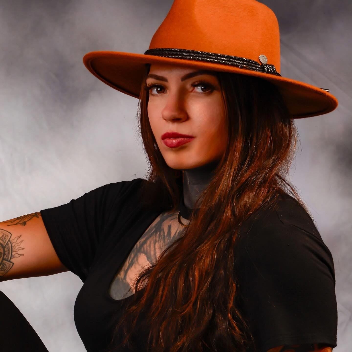 A woman with long red hair wearing a large orange wide-brimmed hat with a black band, a black t-shirt, and tattoos on her arm and chest. She is looking at the camera against a gray background.