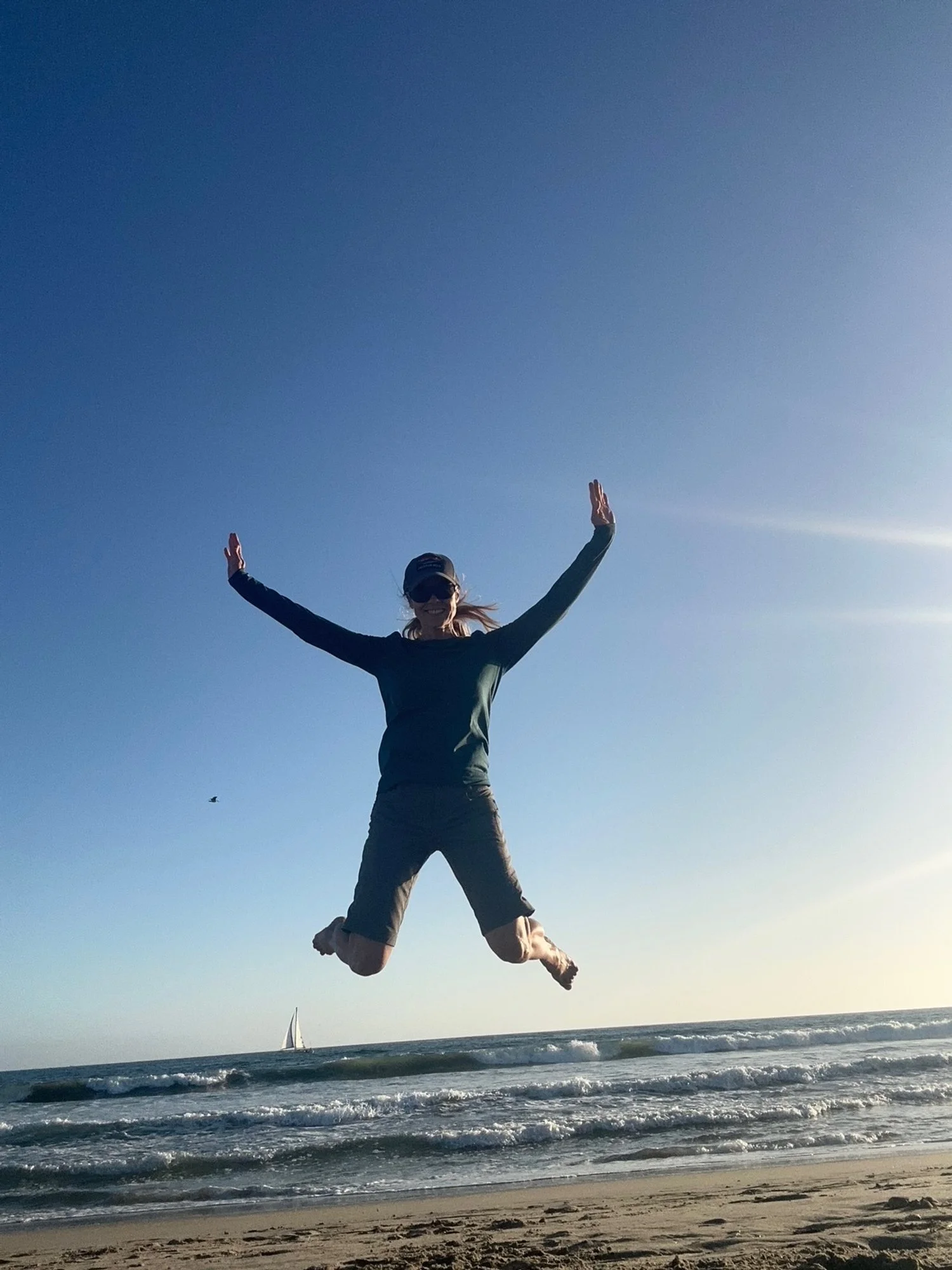 Person jumping on the beach with arms and legs spread wide, blue sky in the background, sailboat on the water, ocean waves, sandy beach.