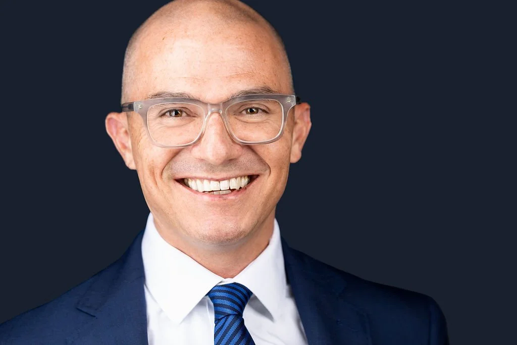 A smiling man in a navy blazer, white shirt, and a blue striped tie, wearing clear glasses, against a dark background.