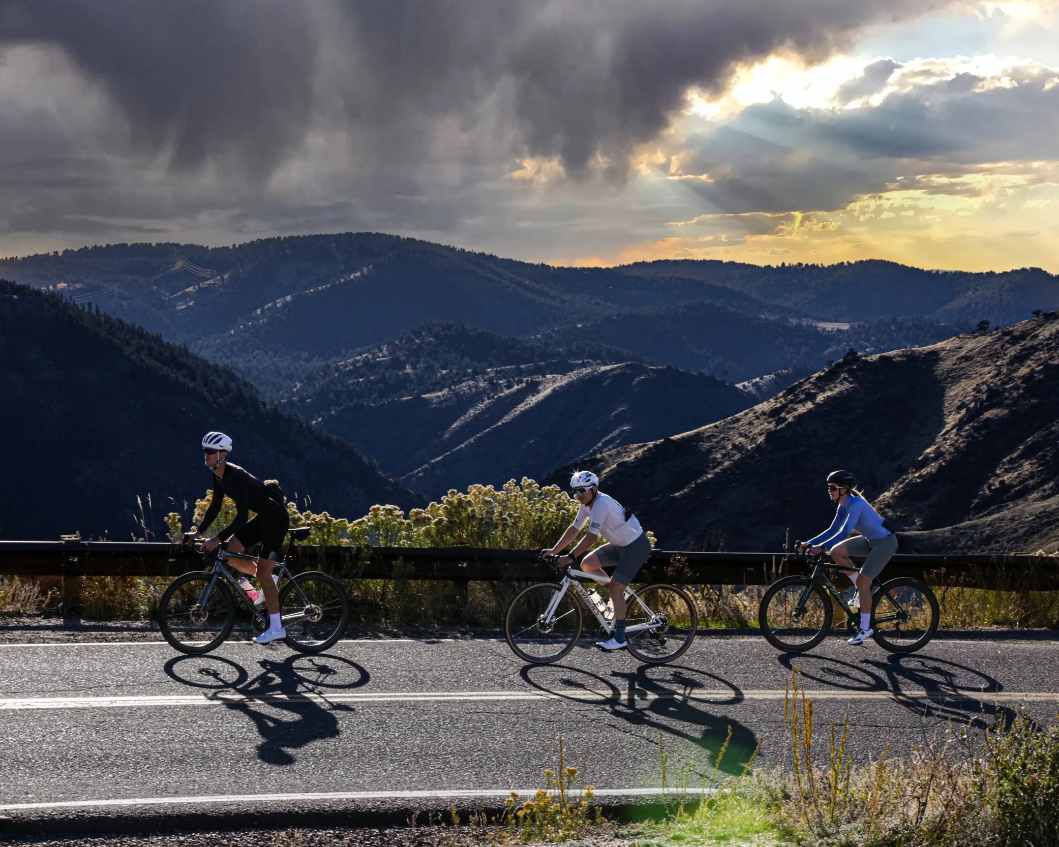 Three cyclists riding on a mountain road during sunset with mountains and clouds in the background.