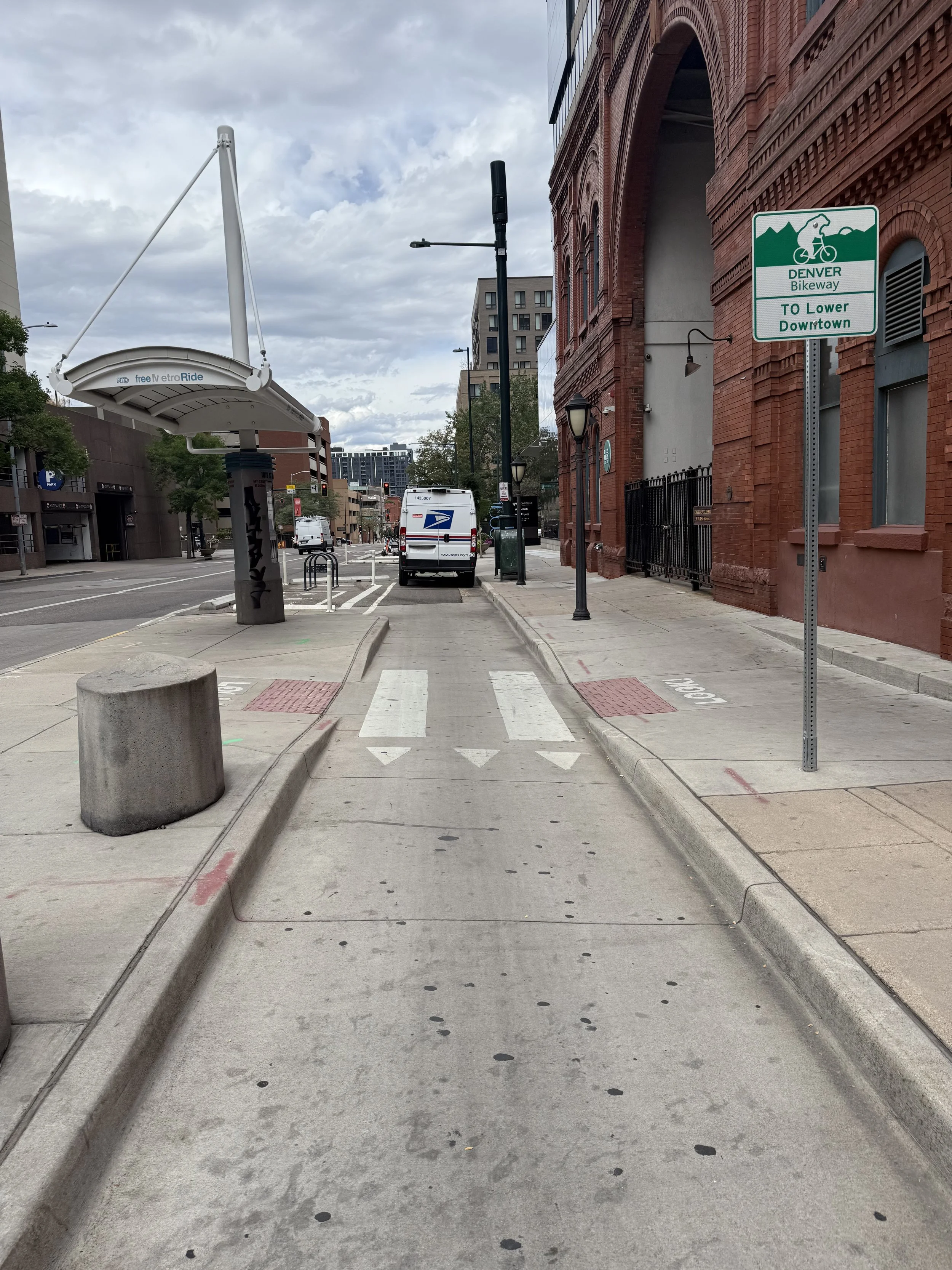 City sidewalk with a designated bike lane, a sign indicating Denver Bikeway to lower downtown, a postal service truck parked nearby, and buildings with cloudy sky in the background.