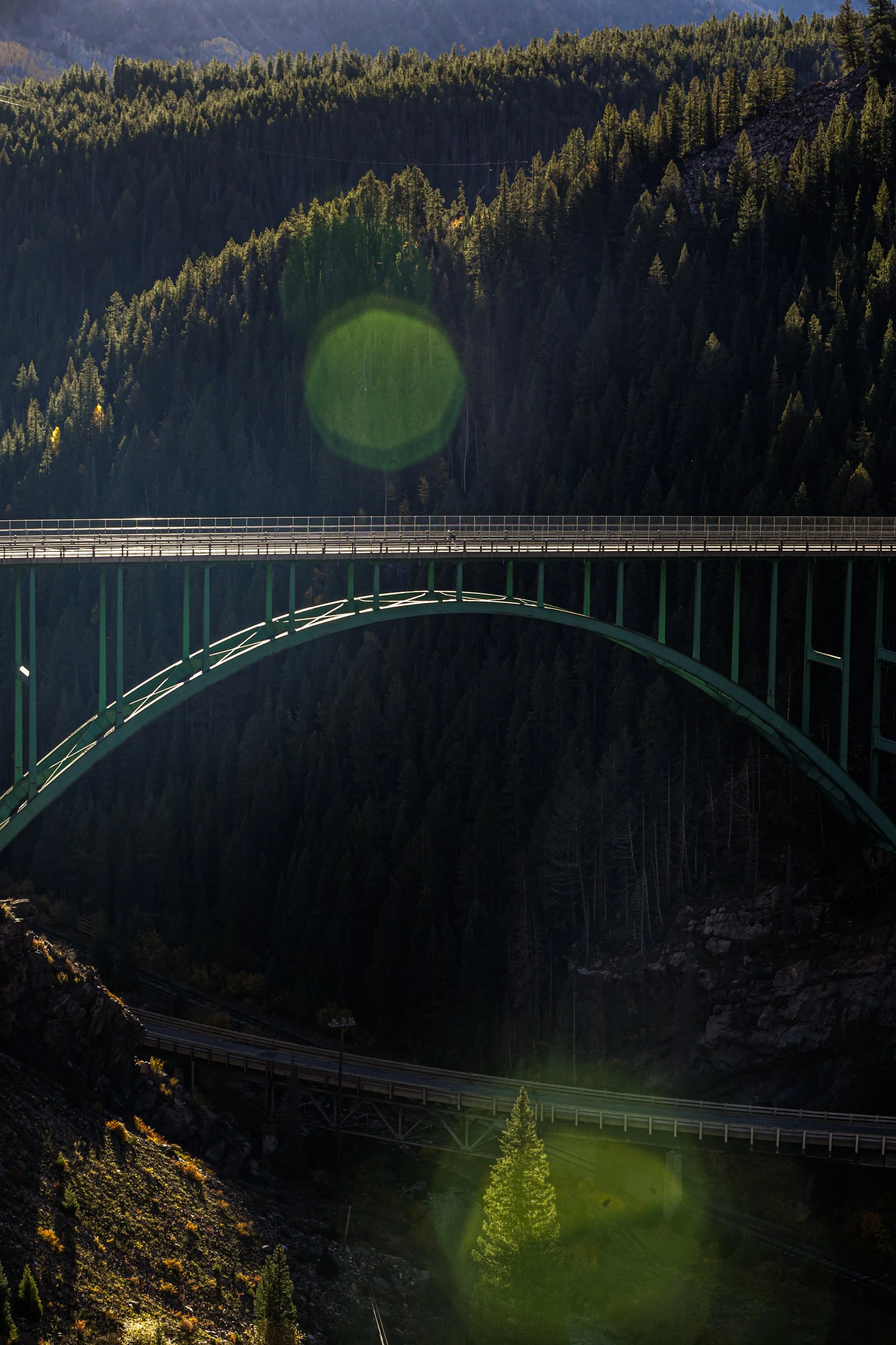 A green arched bridge spanning over a deep forested valley with hills covered in pine trees, and a mountain range in the background.