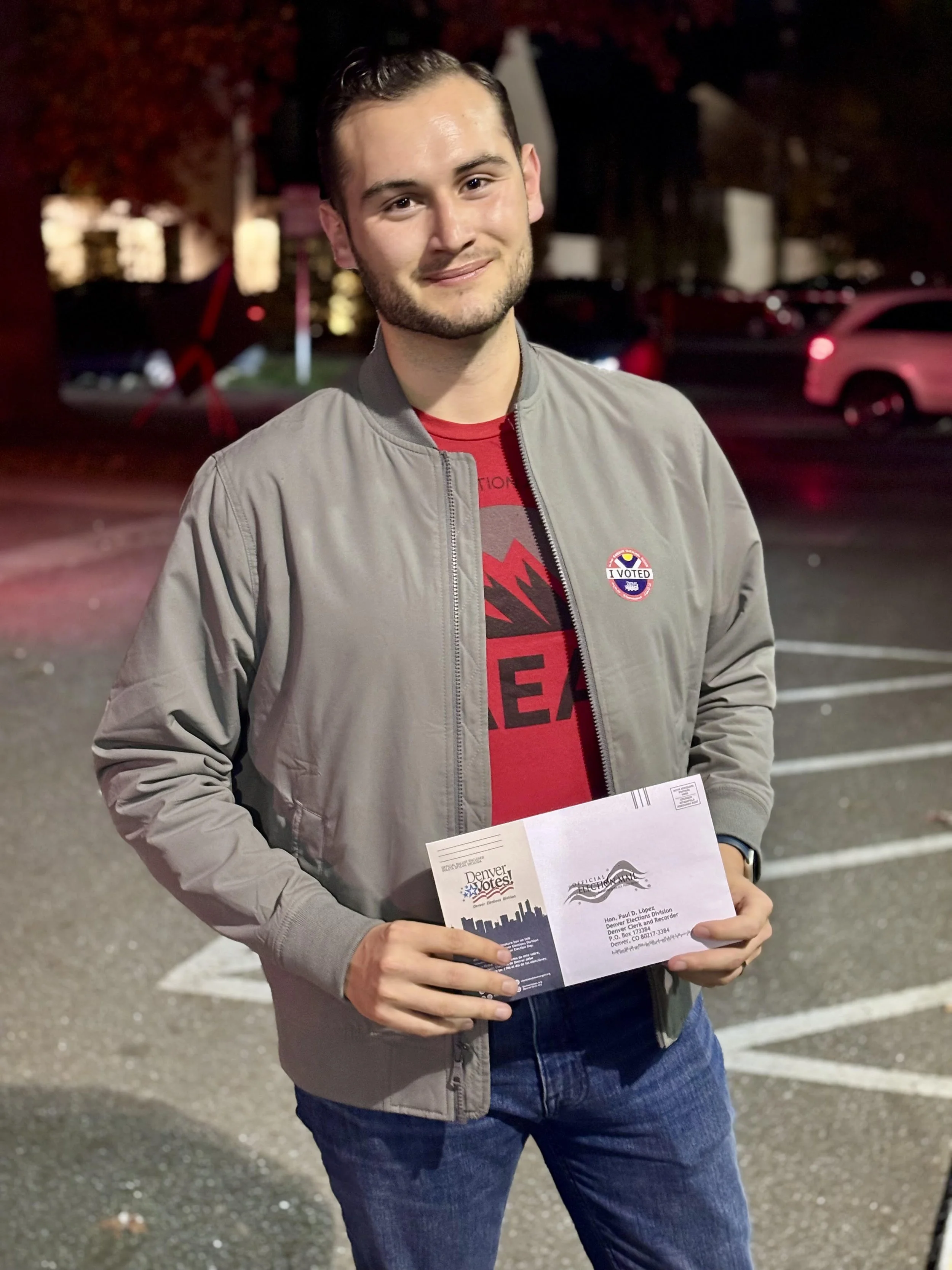 Santiago Palomino standing in a parking lot at night, holding a voting ballot envelope, wearing a grey jacket with a voting sticker, smiling at the camera.