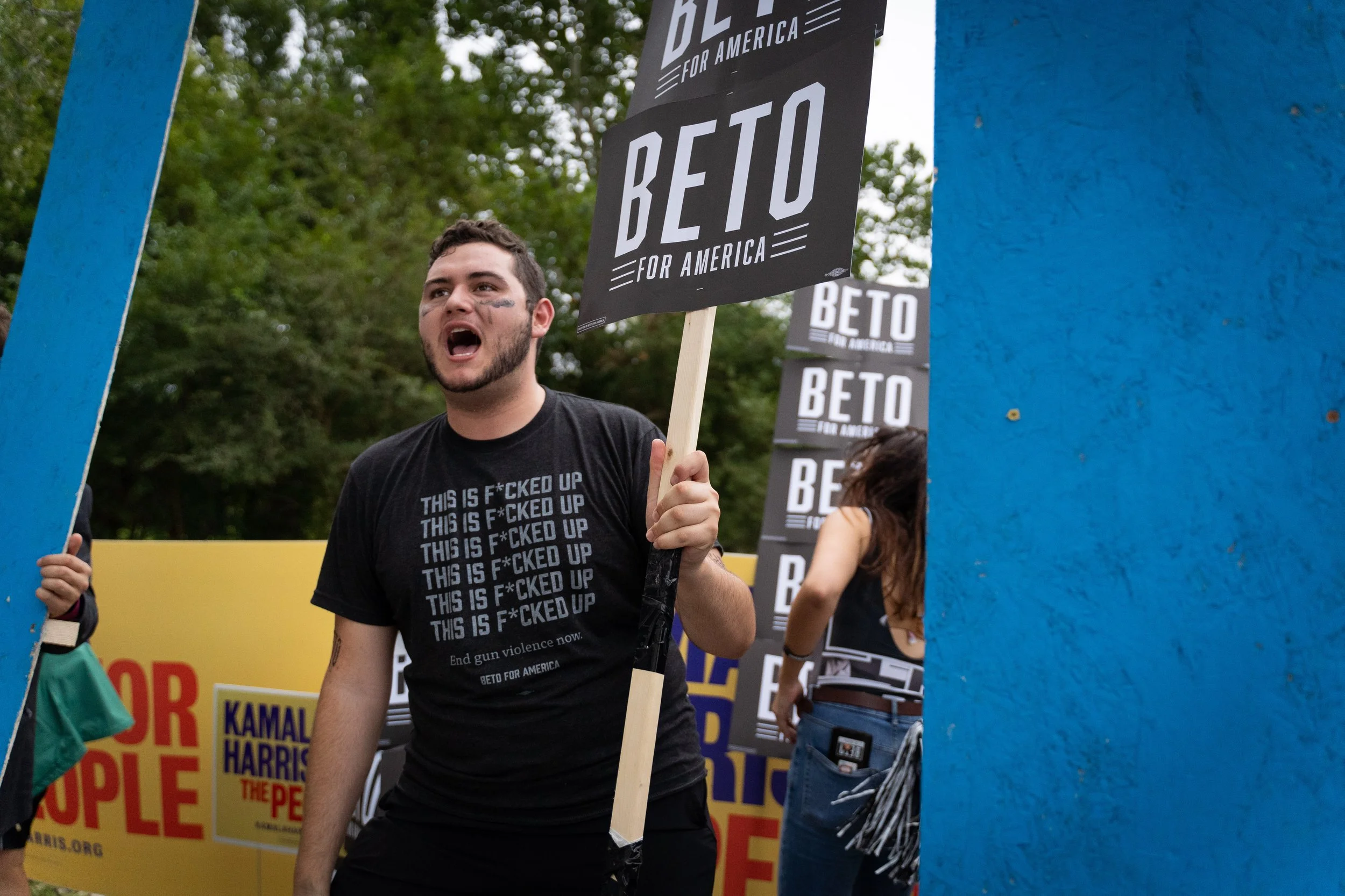 Santiago Palomino protesting at a rally, holding a sign that says 'BETO FOR AMERICA', with other protesters and campaign signs in the background.