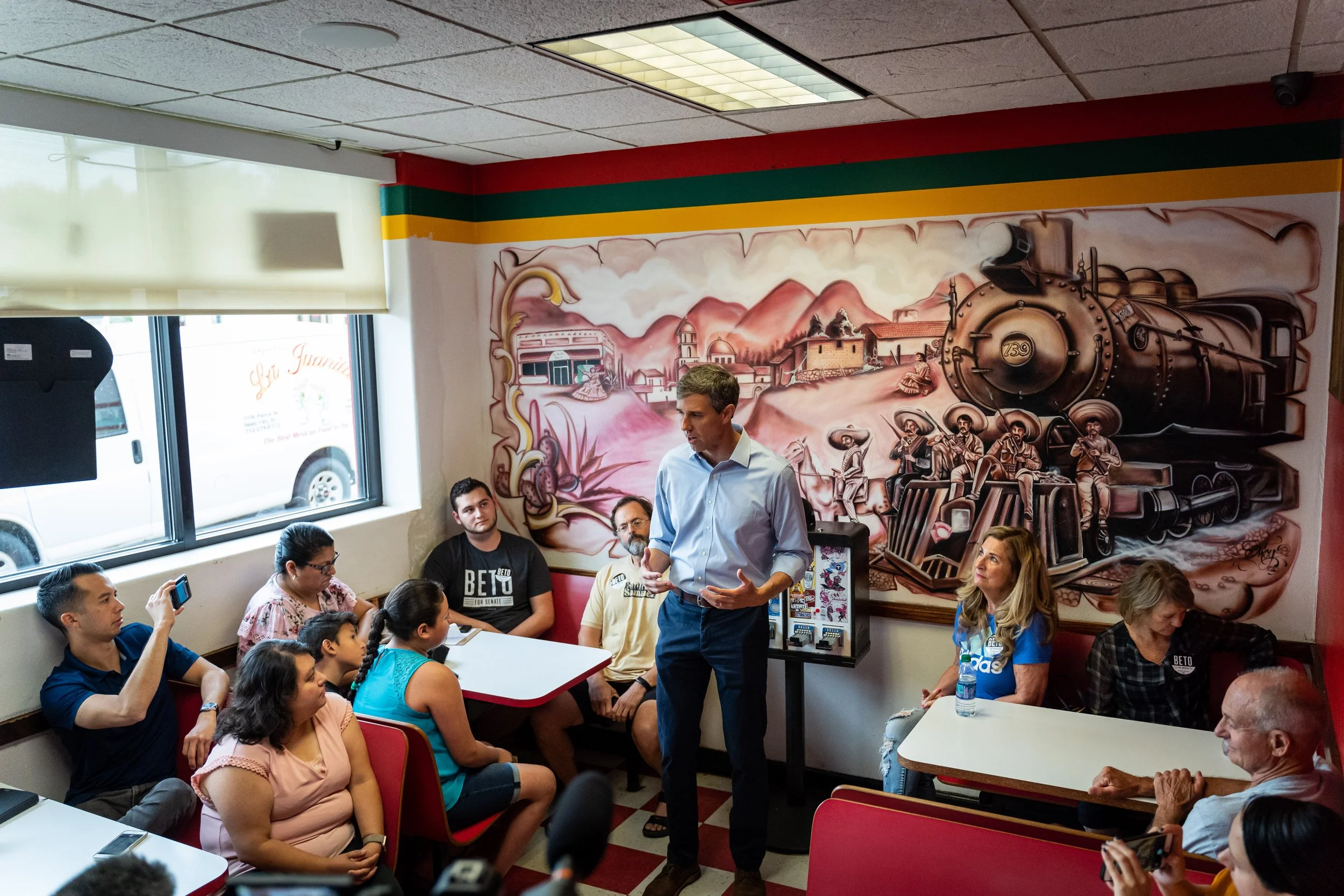 Beto O'rourke in light blue shirt speaking to seated diverse group including Santiago Palomino in a room with a train mural on the wall.