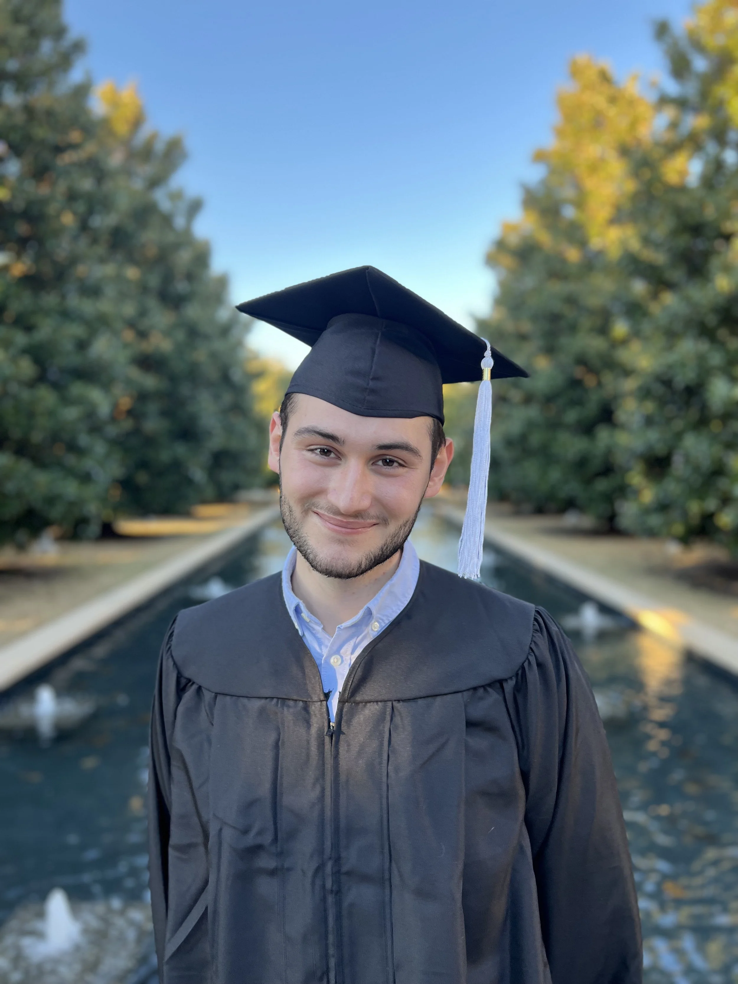 Santiago Palomino wearing a graduation cap and gown, smiling, standing outdoors near a fountain with trees and blue sky in the background.