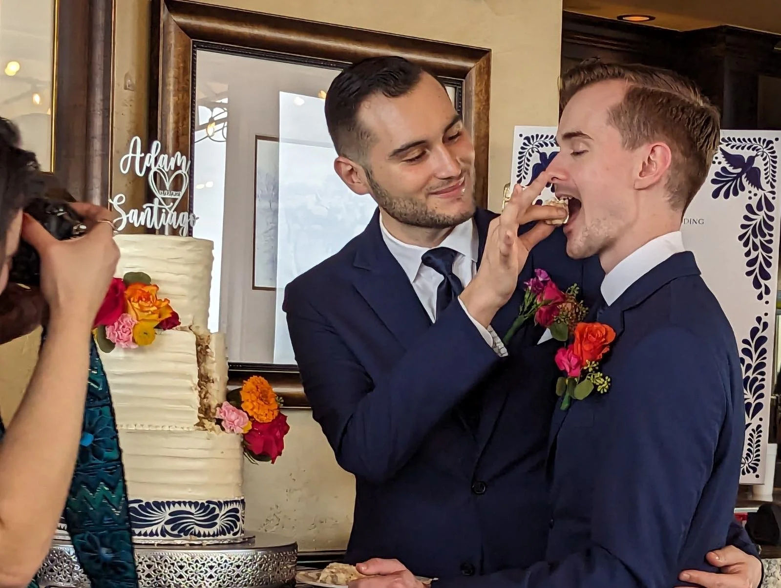 Santiago Palomino and his husband in suits, likely at a wedding, sharing a piece of cake as one helps the other eat it. They are smiling and embracing, with a decorated wedding cake and flowers in the background.
