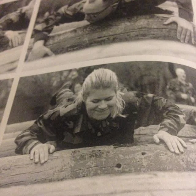 Santiago Palomino's mother in military camouflage leaning over a wooden log, smiling.