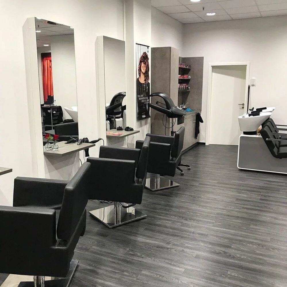 Ein Friseursalon mit schwarzen Friseurstühlen, Spiegeln und Haarwaschbecken. Hinweise auf Haarschnitte und Pflege.
