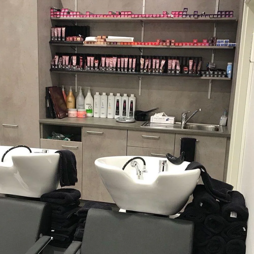 Friseursalon mit Haarkosmetikprodukten und zwei Waschbecken für Haare. Im Hintergrund sind Regale mit Haarfärbemitteln und weiteren Pflegeprodukten.