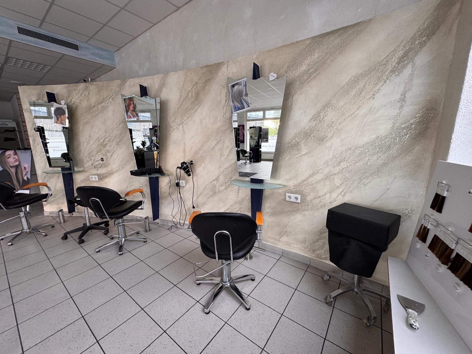 Friseursalons mit drei Spiegeln, Haarschneideplätzen, Kundenstühlen, und Kamm- und Farbproben an der Wand, im Hintergrund Fenster mit natürlichem Licht.