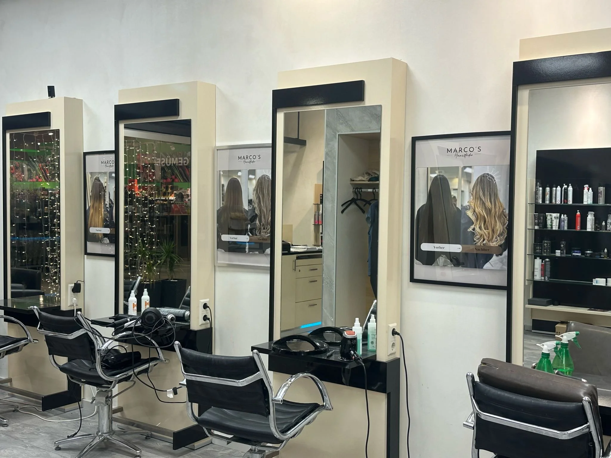 Friseurstühle vor Spiegeln in einem Friseursalon, mit Haartrocknern und Pflegeprodukten auf den schwarzen Tischen.
