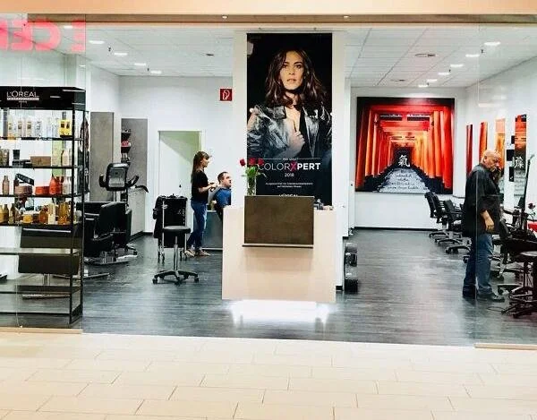 Innenansicht eines Friseursalons mit Kunden und Friseuren, ausgestatteten Spiegeln und Stühlen, Wandbildern, und einem Empfangstresen im Vordergrund.