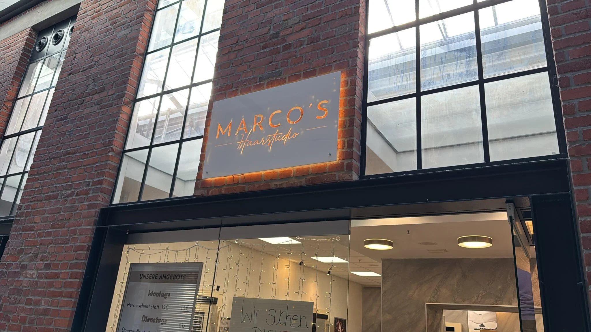 Das Bild zeigt den Eingangsbereich eines Friseursalons namens 'Marco's Haarstudio' in einem Gebäude mit Backsteinfassade und großen Fenstern. Es sind Lichter und ein Willkommensschild im Fenster sichtbar.