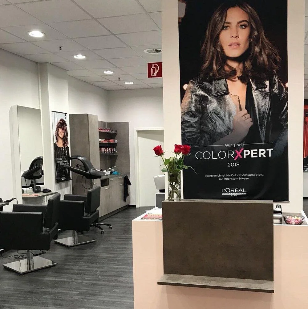 Innenraum eines Friseursalons mit schwarzen Stühlen, Waschanlagen, einem Regal mit Produkten und einem großen Poster einer Frau mit lockigem Haar und Lederjacke. Auf einem Tisch steht eine Vase mit drei roten Rosen.