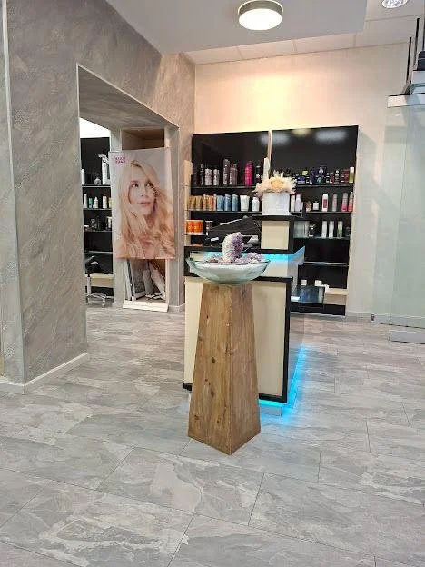 Ein moderner Salon mit einem runden Wassertrog auf einem Holzhocker in der Mitte. Im Hintergrund sind Regale mit Haarpflegeprodukten und ein Poster einer Frau mit langen, gewellten blonden Haaren. Die Räume sind hell und stilvoll gestaltet.