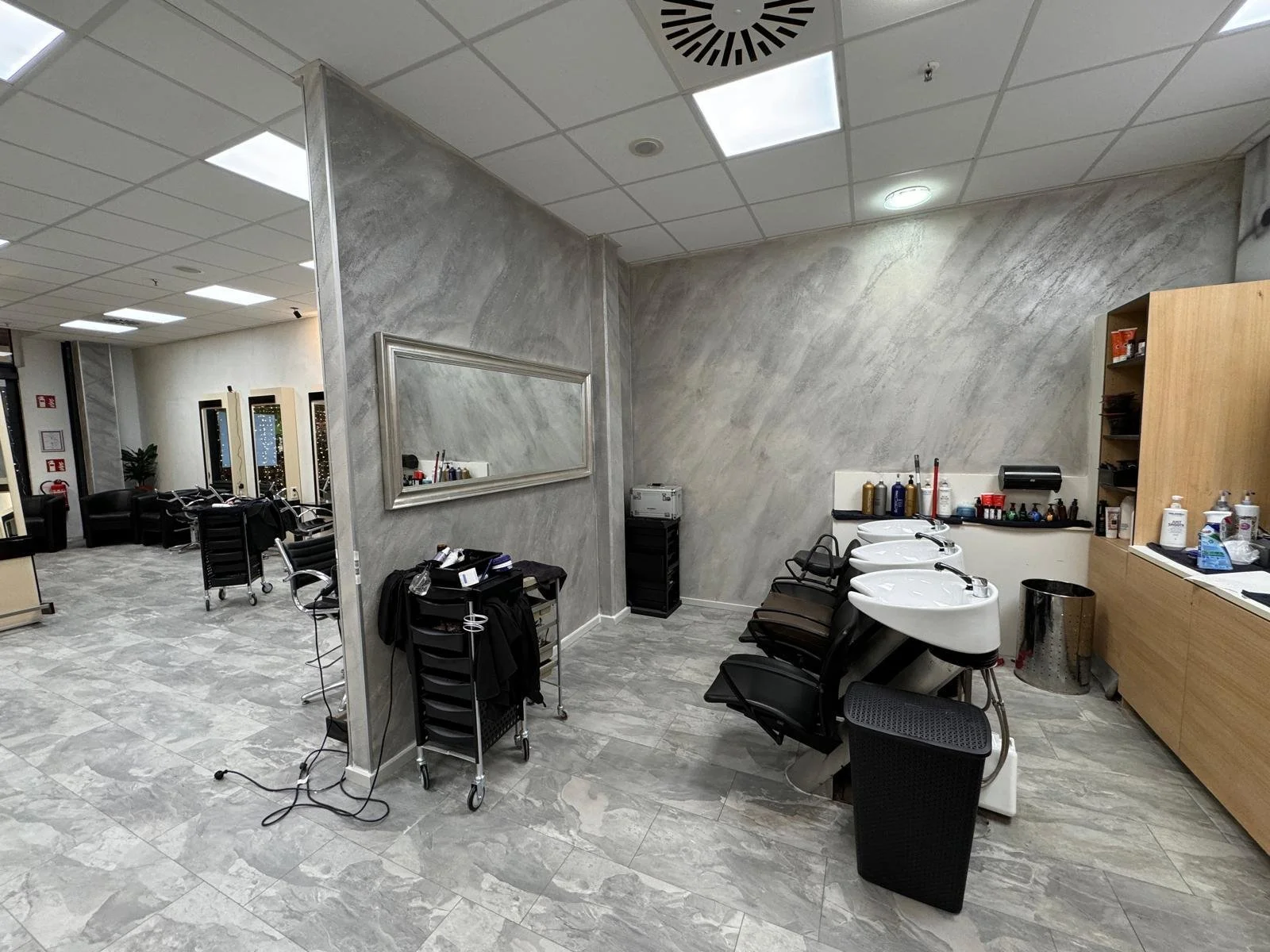Friseursalon mit vier Waschbecken, schwarzem Stühlen, Holzwand und kleinen Regalen mit Haarprodukten. Sitzbereich mit schwarzen Sesseln im Hintergrund.