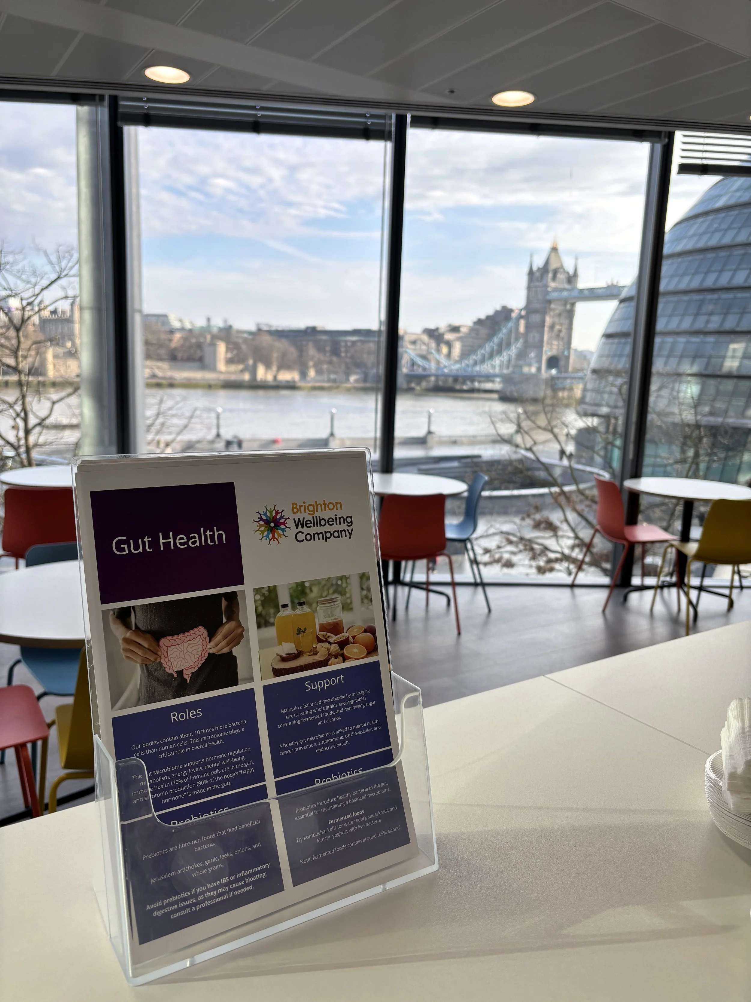 Indoor cafe with view of Tower Bridge in London through large windows, colorful chairs, and a table with a pamphlet about gut health.