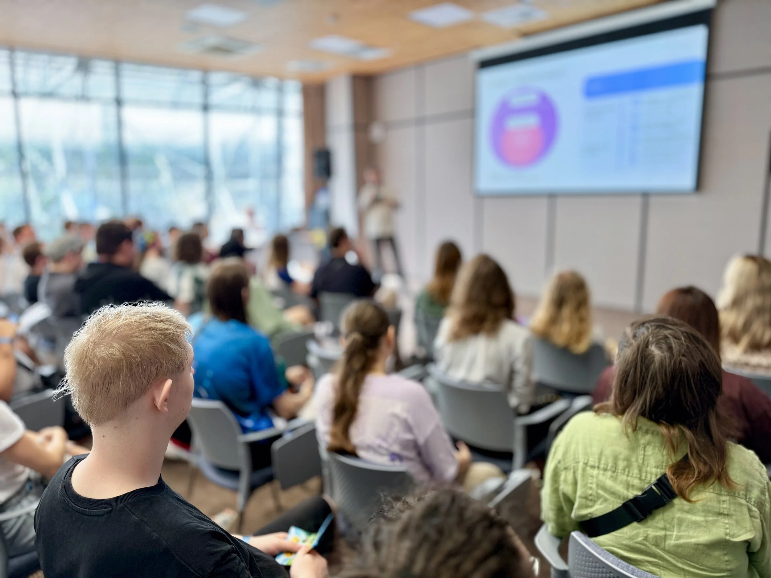A group of people seated in an auditorium watching a presentation on a large screen.