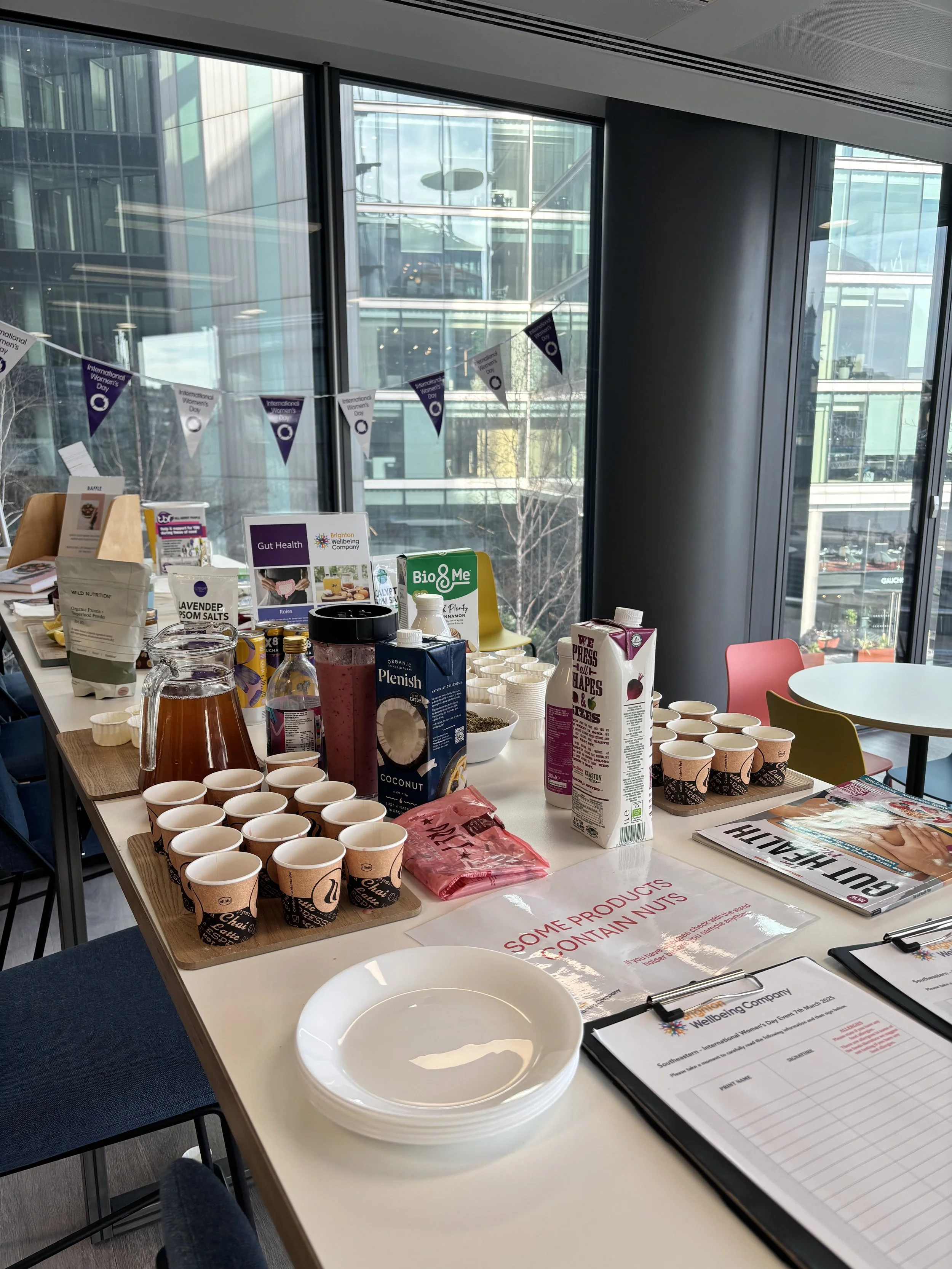 Table with various food and drink items, paper cups, clipboards, and informational signs set up for an event in a modern office or conference room with floor-to-ceiling windows.