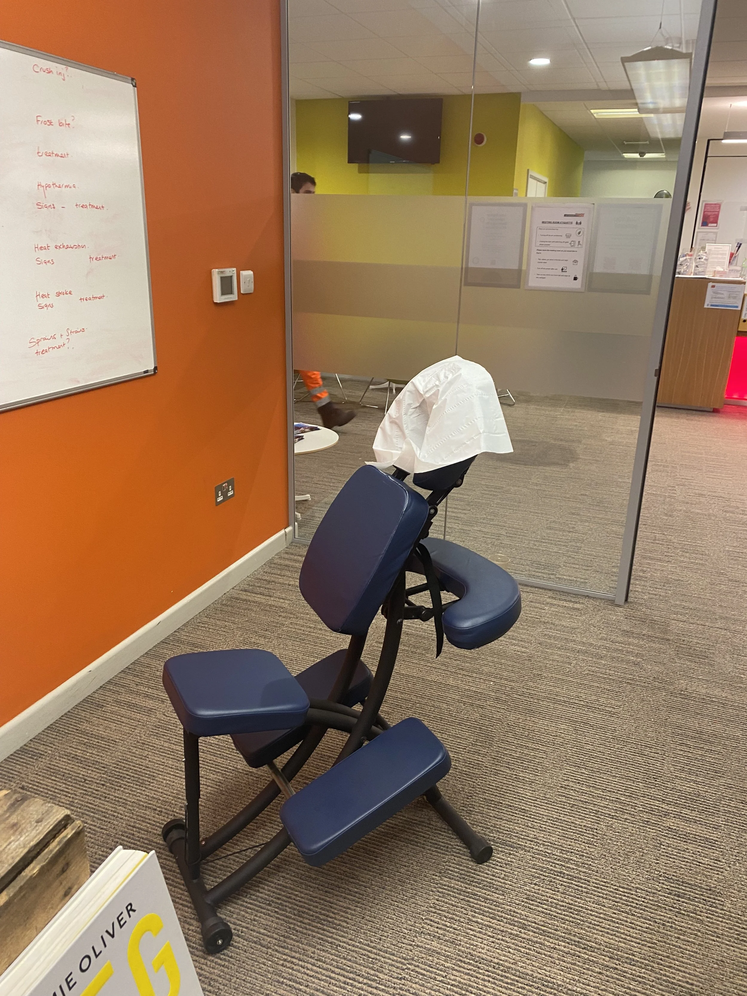 An exercise chair with a white cloth draped over one of the backrests is positioned in front of a glass wall in an office or medical clinic. Behind the glass are yellow and green painted walls, a mounted TV, and several documents posted on the wall. 