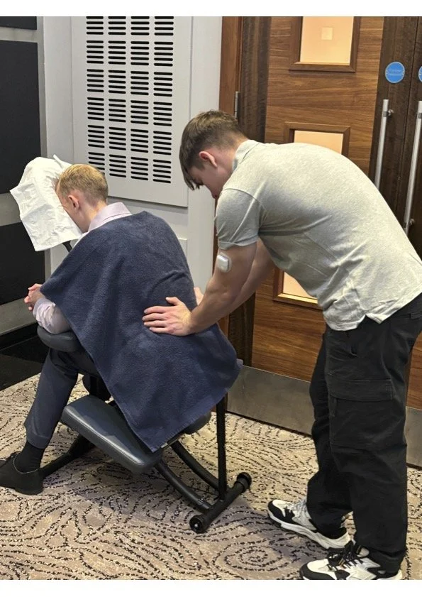 A person with a medical device on their arm is leaning forward in a chair with a towel draped over their shoulders, while another person is providing a massage or physical therapy in a room with wooden walls and a patterned carpet.