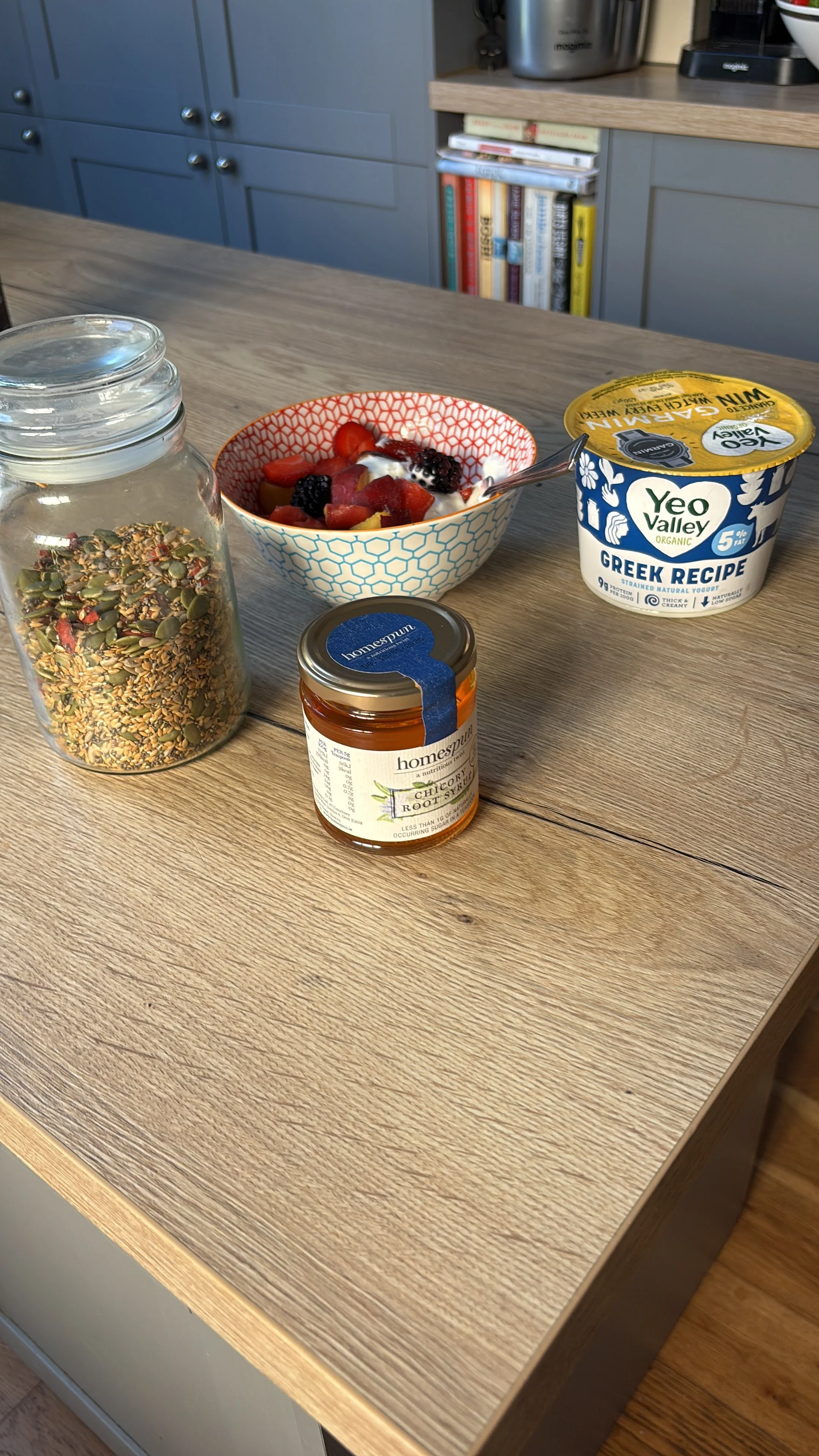 A wooden kitchen table with a jar of granola, a bowl of mixed berries and yogurt, a container of Greek yogurt, and a jar of honey.