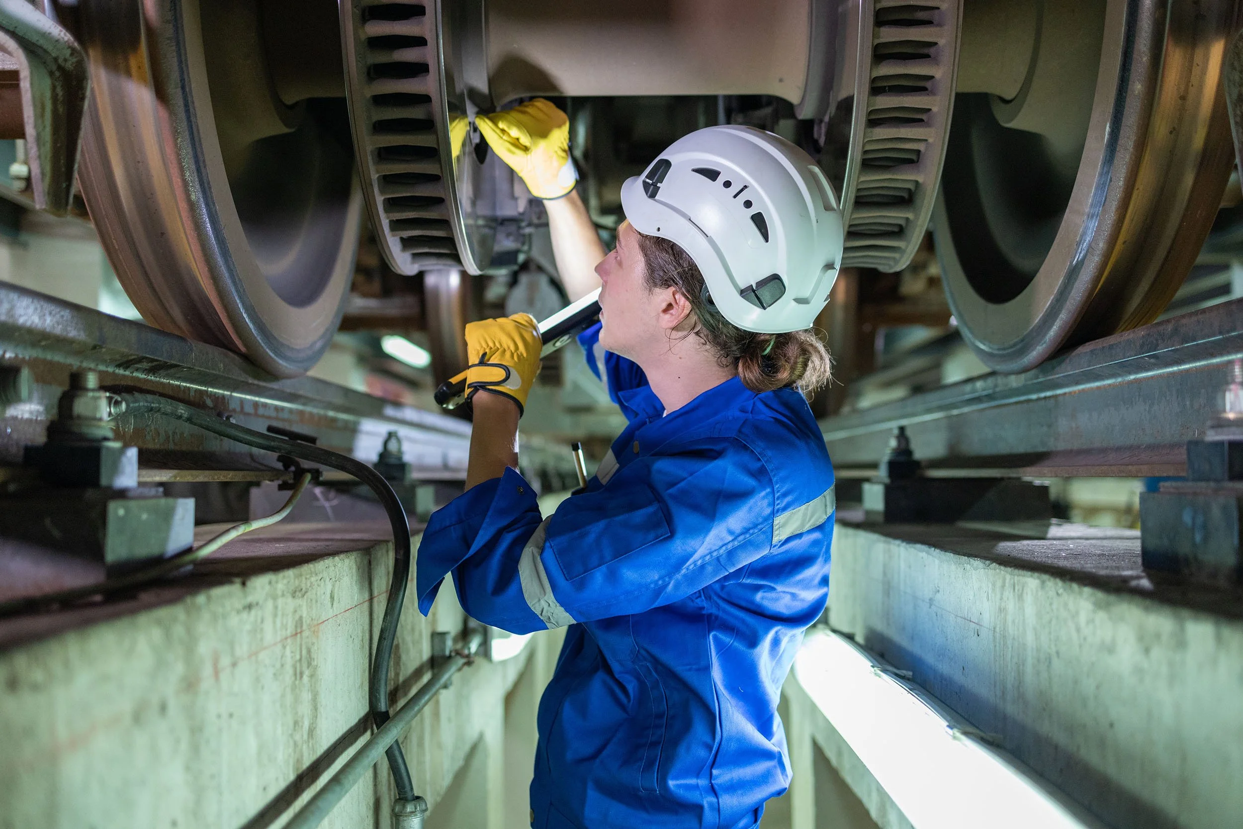 A worker wearing a white helmet and blue uniform inspecting or repairing a large industrial machine with big rollers.