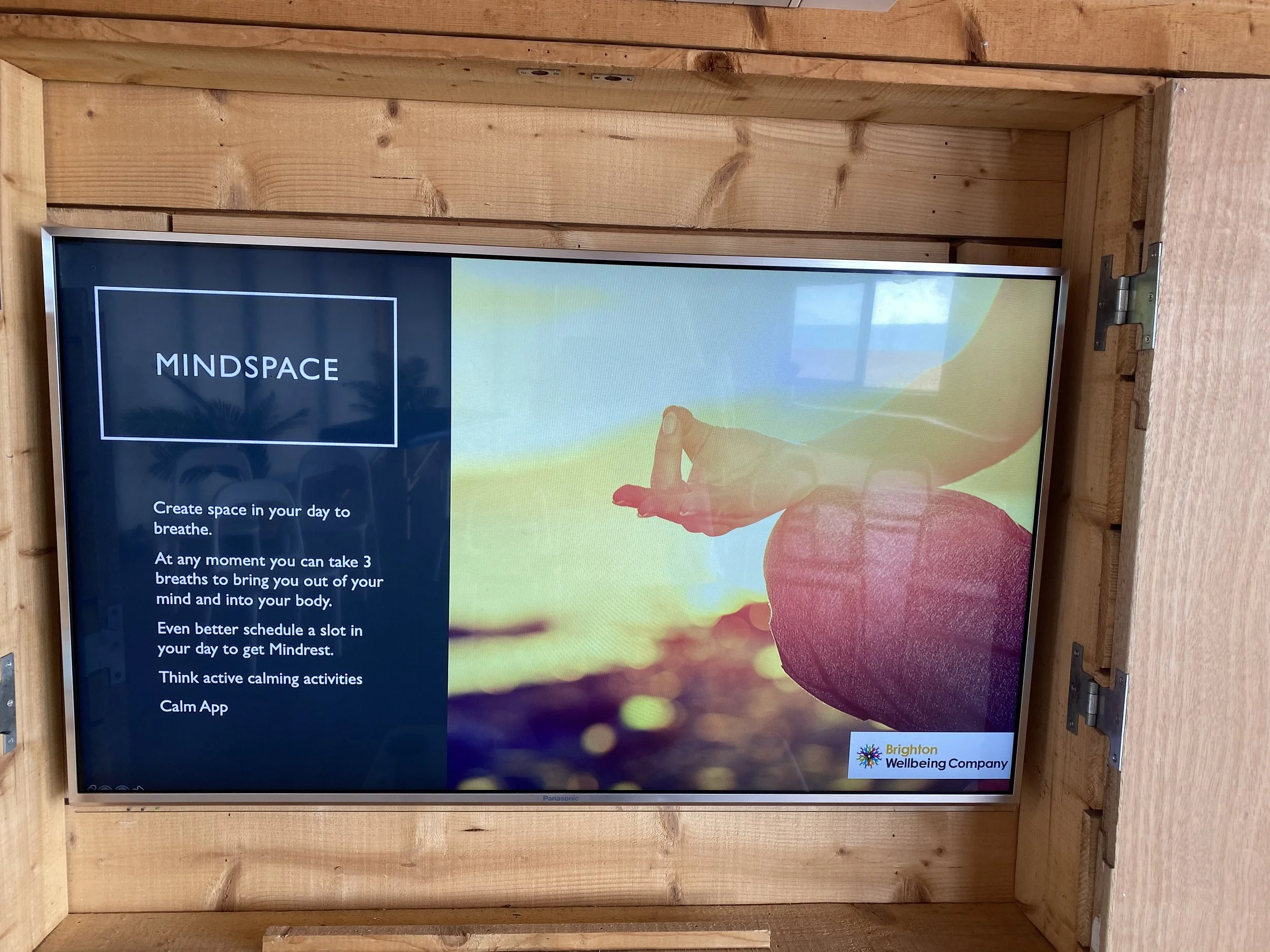 A digital information screen about mindfulness in a wooden frame. The screen shows a person in a yoga pose holding their fingers in a mudra, with a sunrise or sunset background. The left side of the screen has text about creating space in your day to